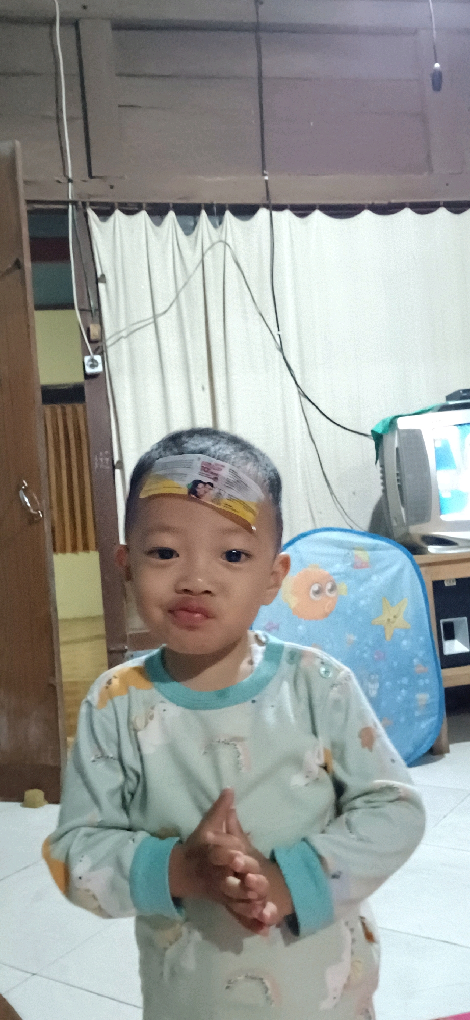 A young boy with a sticker on his forehead stands in a room. He is wearing a light blue long-sleeved shirt with cartoon animal prints. His hands are clasped in front of him. In the background, there is a white curtain, a wooden door, a television set, and a blue laundry basket with cartoon fish and stars on it. The floor is tiled. The image appears to be taken indoors, possibly in a residential setting in Wonokerso, Indonesia.