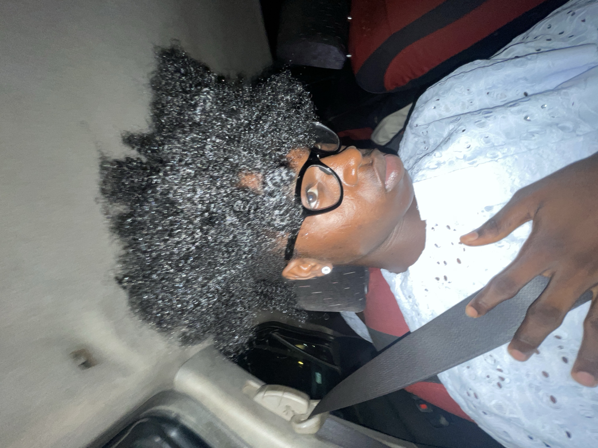 The image shows a woman in a car, wearing glasses, a white patterned shirt, and a seatbelt. The car interior has a dark red and black striped seat cover. The woman has dark skin and curly black hair. The location is indicated to be Igboefon, Nigeria, however, there are no visual clues in the image to independently verify this location.