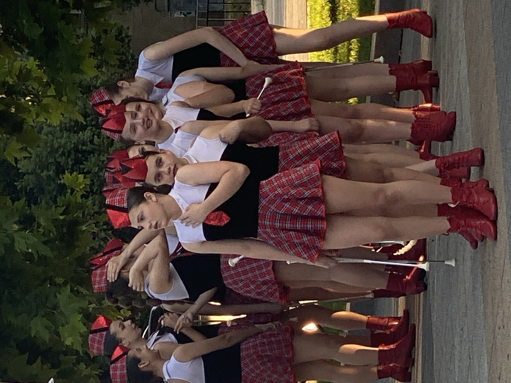 The image shows a group of young women, likely a dance or baton twirling troupe, dressed in matching red plaid skirts, black tops, and red boots. They are arranged in a staggered line, appearing to be in motion or posing for a performance. Each of them holds a white baton. The women have serious expressions, suggesting focus and concentration on their activity. The scene is outdoors, with green foliage visible in the background and a paved surface beneath them. The lighting suggests it is daytime, possibly late afternoon or early evening, as indicated by the warm light. The orientation of the image is vertical. The backdrop with trees and paved ground might indicate a park, school grounds, or a public square in Pleven, Bulgaria. The overall impression is one of a synchronized performance or rehearsal. There is no visible text in the image.