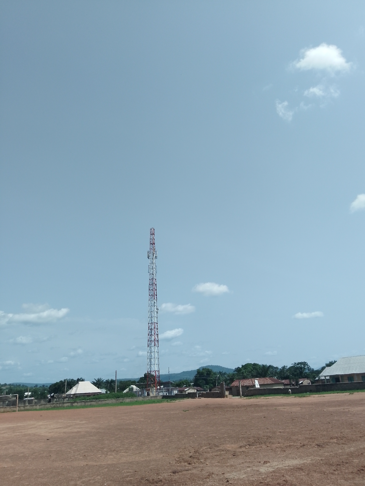The image depicts a wide, open, dirt field in the foreground, likely a sports ground, with a faint outline of a goalpost visible on the far left. Beyond the field, a low, weathered brick wall stretches across the mid-ground, marking the boundary of a settlement. Several buildings with corrugated metal roofs and some with whitewashed walls are visible behind the wall, interspersed with lush green trees and palm trees. Dominating the background is a tall, red and white telecommunications tower, standing prominently against a pale blue sky dotted with scattered white clouds. In the far distance, rolling hills form the horizon. The scene appears to be set in the town of Serti, Nigeria. There are no people visible in the image.