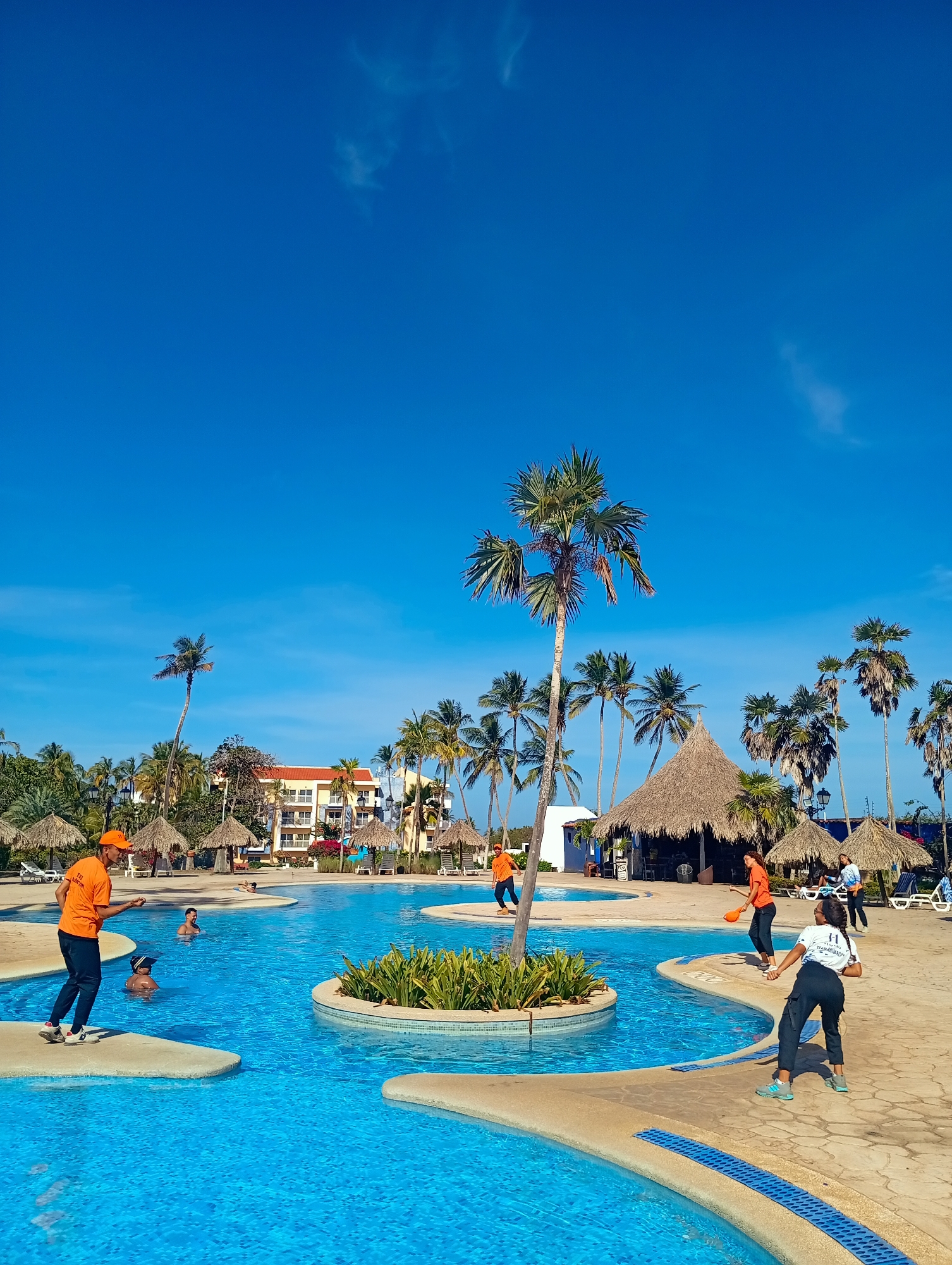 A sunny day at a resort in Playa El Agua, Venezuela. A large blue swimming pool, surrounded by palm trees and thatched-roof structures, occupies the center. Several people are present. A man in an orange shirt and hat is positioned on the left edge of the pool, appearing to be throwing something. A woman in a white shirt and black pants is on the right edge, also seemingly preparing to throw an object. More people are in the water, and one individual is mid-stride near the center of the pool, also clad in orange. The sky is bright blue with a few scattered clouds. In the background, there's a multi-story building. Lounge chairs are visible near the thatched roofs.