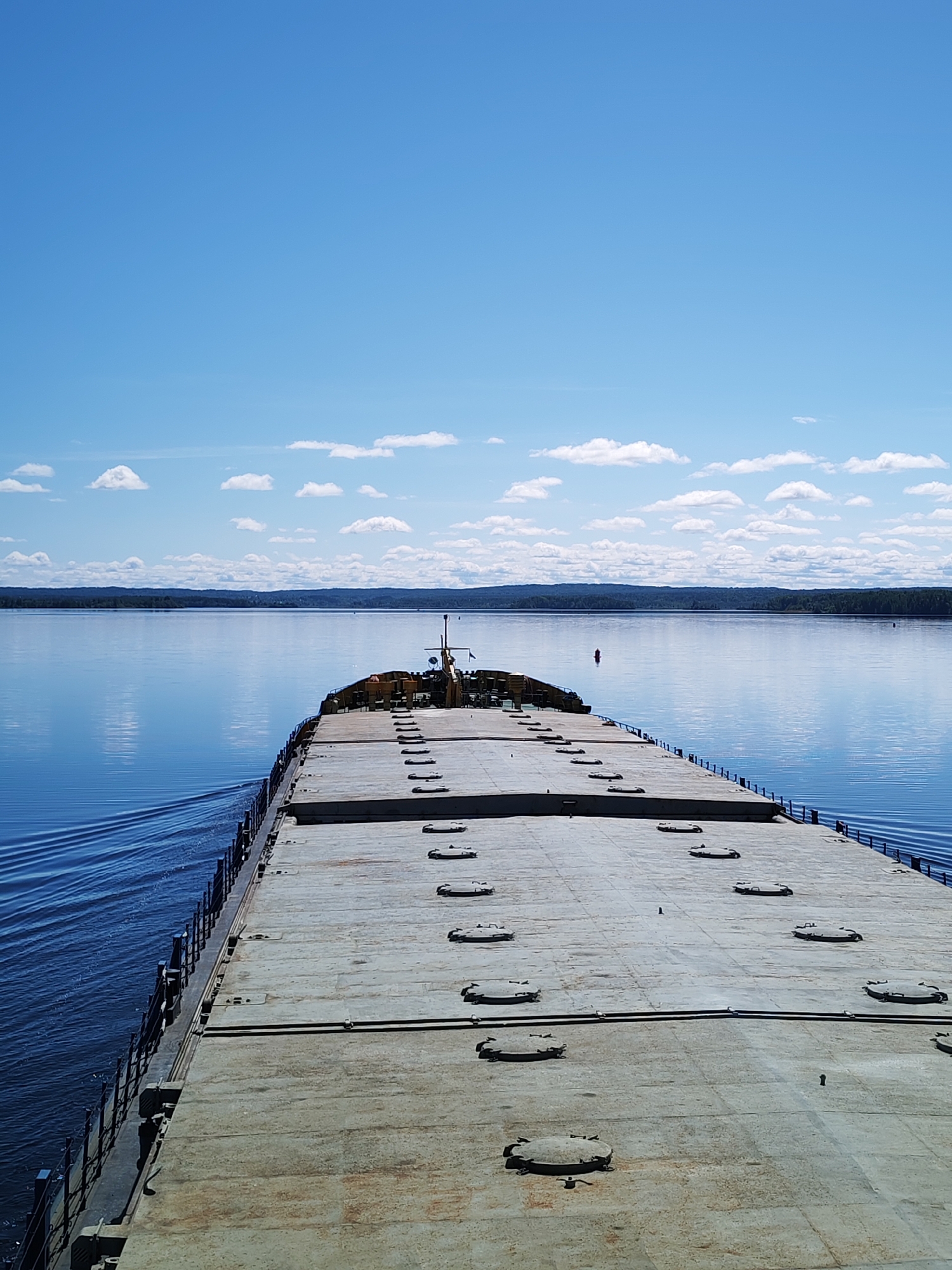 This image presents a serene daytime view from the deck of a large vessel, likely a barge or cargo ship, navigating a wide body of water near Shestovo, Russia. The perspective is looking forward over the vessel's long, flat deck towards the horizon.
The foreground is dominated by the ship's deck, a broad, weathered expanse of gray or off-white metal. Numerous circular hatches, possibly covers for cargo holds, are evenly spaced across the deck. Towards the bow, some industrial equipment or a small superstructure is partially visible, along with railings lining the sides of the ship.
To the left of the vessel, a distinct wake trails across the calm, deep blue water, indicating the ship is moving forward. The water beautifully reflects the bright blue sky above, creating a seamless blend of blues. A single red buoy can be seen floating in the water in the middle distance, to the right.
In the far distance, a low, dark green tree-lined horizon stretches across the frame, marking a distant shoreline or landmass. The sky above is a clear, vibrant blue, adorned with scattered, fluffy white cumulus clouds.
The scene conveys a sense of peaceful travel under clear, sunny weather conditions, likely mid-morning or afternoon given the bright illumination. No people or visible text are present in the image.