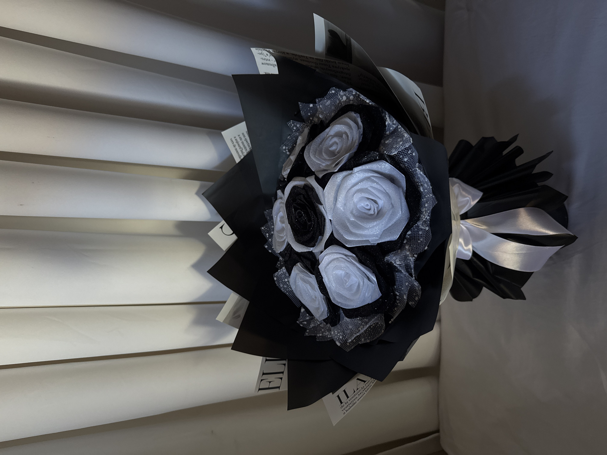 The image displays a stylish bouquet of artificial roses, predominantly in black and white/silver tones, arranged in an indoor setting. The bouquet features several white/silver roses with a glittery finish and some black roses, also with a subtle shimmer. These flowers are encased in layers of black wrapping paper, interspersed with pages from a magazine or book that serve as part of the decorative wrapping. Visible text on these pages includes phrases in French such as "de la couture", "la rue", "vestiaires", and "a aujourd'hui", alongside Polish text mentioning "Shaw, dzis", "fotograficznych", "stylist", "albumow", and "YouTube". A white satin ribbon is tied around the base of the bouquet, forming a neat bow. The bouquet is positioned against a background of light-colored, horizontal slatted or paneled surfaces on the left, possibly blinds or a wall feature, and a darker, plain surface on the right. The lighting is moderate, creating some shadows and highlighting the glitter on the roses. The overall aesthetic is elegant and modern.