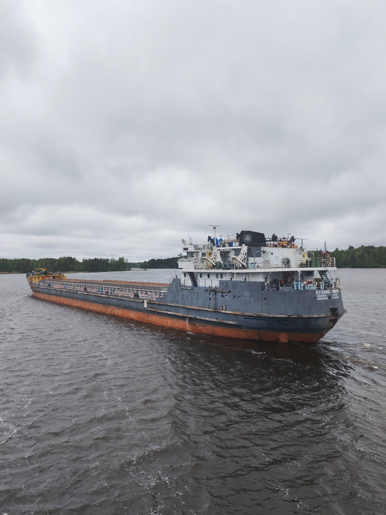 A large cargo ship, named "Kuzma Minin" is sailing on a body of water, with a coastline visible in the background. The ship is grey with an orange lower hull, and has people on the deck. The sky is overcast with grey clouds. The water is dark and choppy. The location is likely Nikol'skaya Gora, Russia, based on the ship's name and common Russian shipbuilding practices.