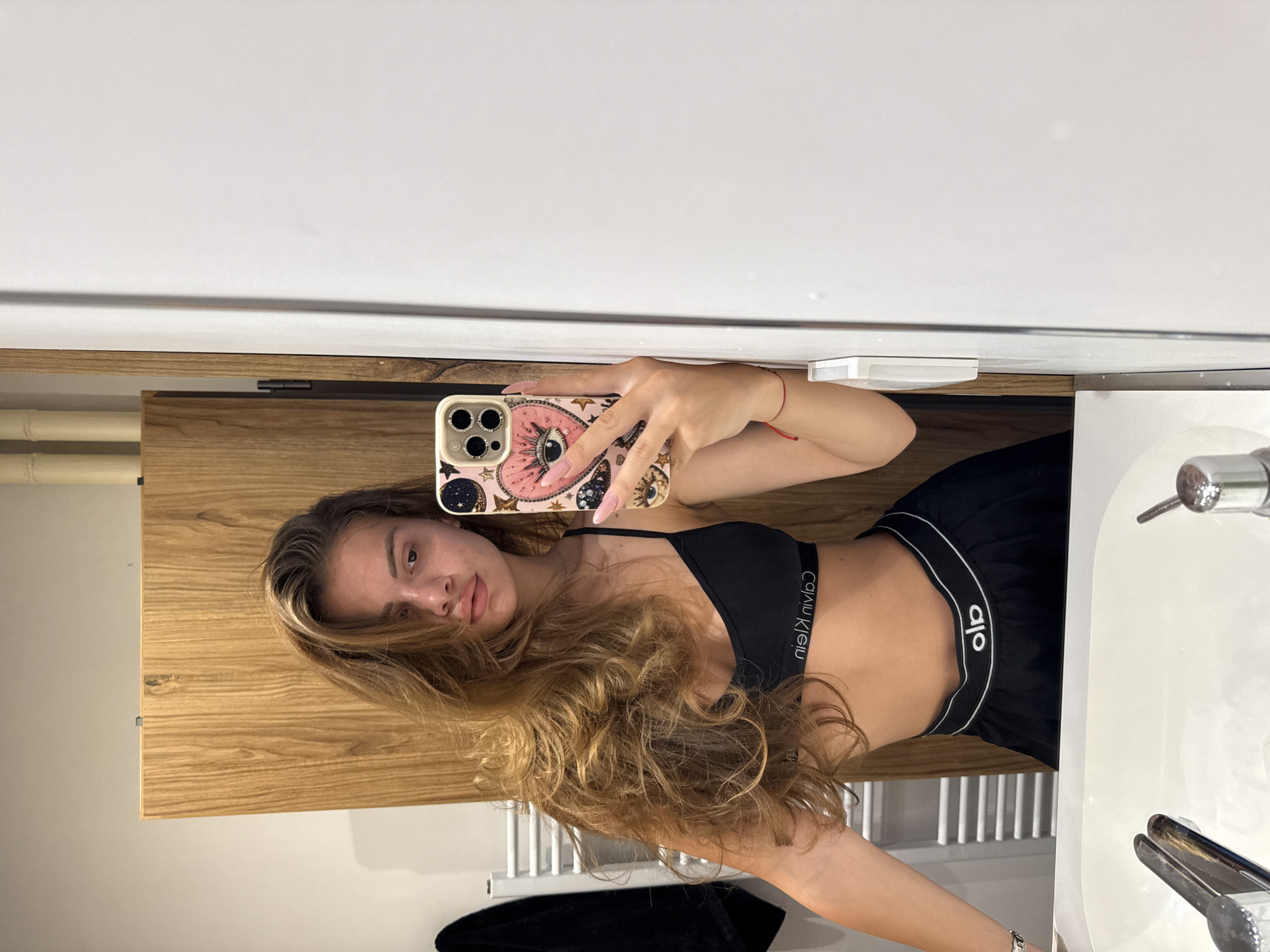 A young woman with long, wavy brown hair is taking a mirror selfie in a bathroom. She is wearing a black Calvin Klein bralette and black shorts with a white waistband. She is holding a pink iPhone with a decorated case in her right hand. A red string bracelet is visible on her left wrist. The bathroom has a wooden cabinet to the left and a white tiled wall. There is a white sink with chrome faucets in the foreground. The scene is lit by artificial light.