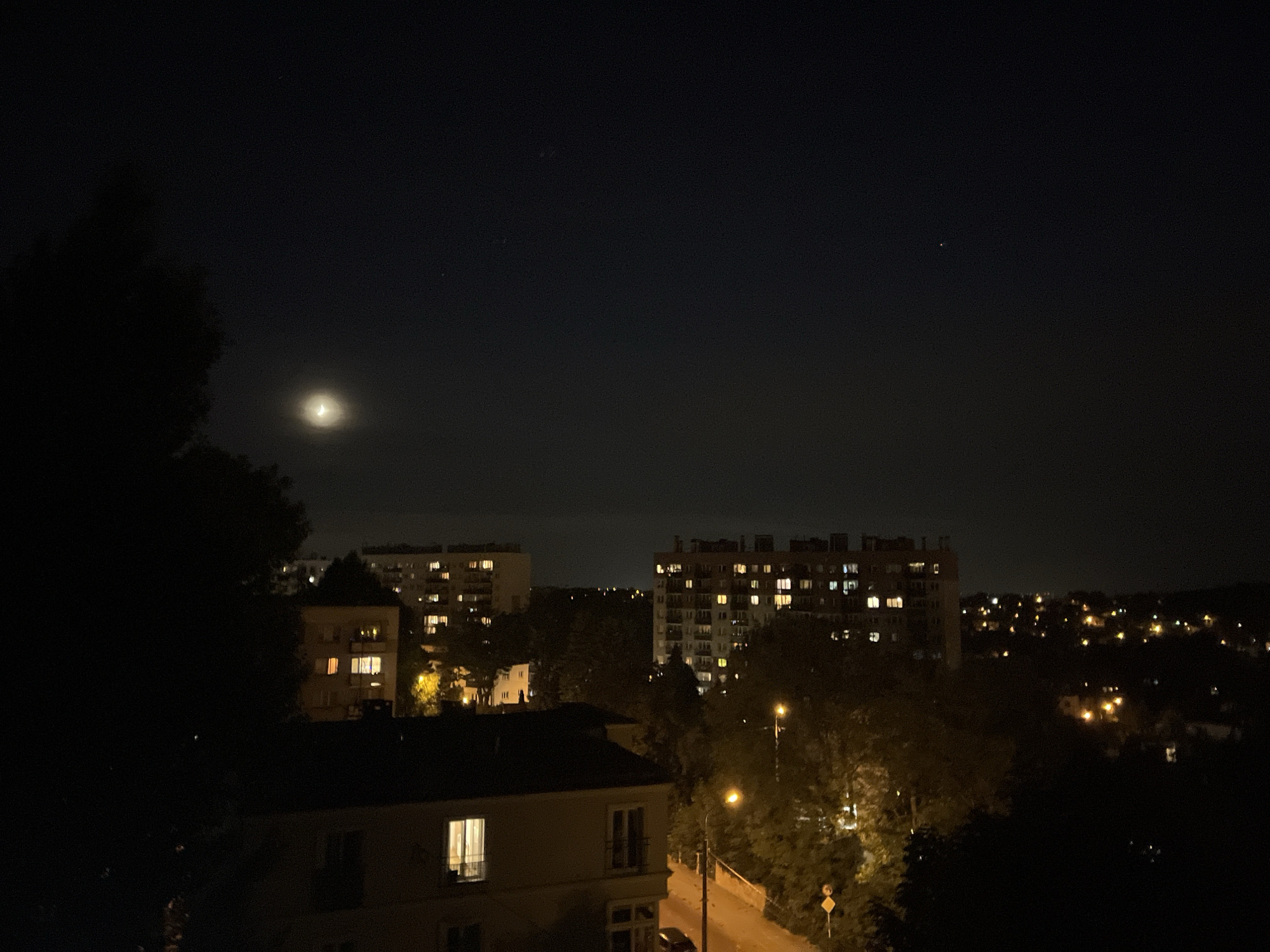 The image shows a night view of a city with buildings and trees illuminated by artificial lights. In the upper left quadrant, a crescent moon is visible in the dark sky. To the right of the moon, there are a few faint stars. In the mid-ground, there are several apartment buildings with lights on in many windows. The buildings vary in height and architectural style. A street lamp with a warm orange glow is visible in the right mid-ground, illuminating a section of a road and surrounding trees. The foreground is dominated by dark foliage, likely from trees, obscuring the lower part of the scene. A small car is parked on the side of the road near the street lamp. The overall impression is a residential area at night, with activity indicated by the lit windows.