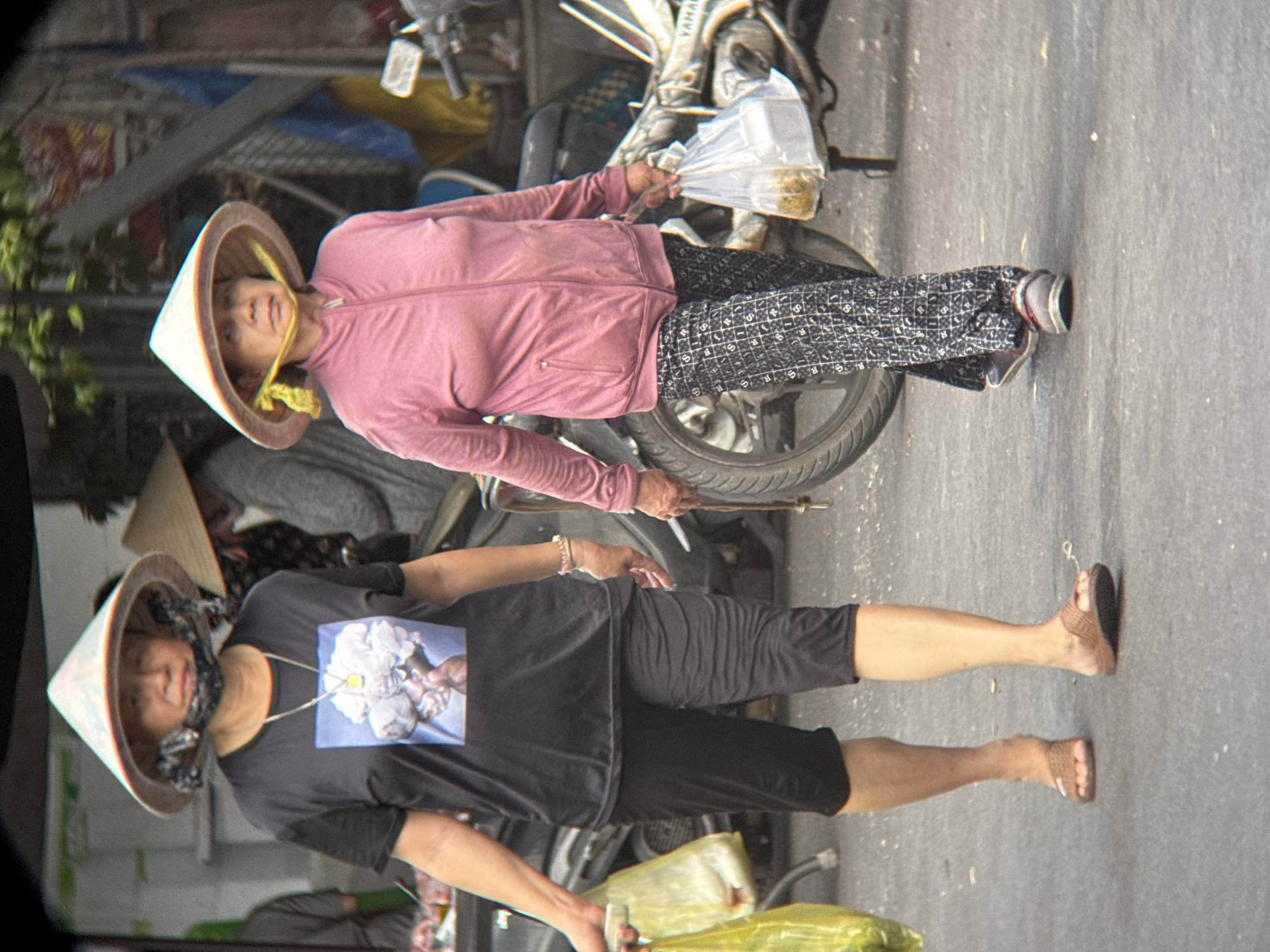 Two women, likely of Vietnamese descent, are walking on an asphalt street in Nha Trang, Vietnam. Both are wearing traditional conical hats (nón lá) and appear to be in their middle ages. The woman on the left is wearing a black t-shirt with a graphic print of a bouquet of white flowers and black pants that appear to be culottes. She is carrying a bright yellow plastic bag in her left hand and wearing brown woven sandals. The woman on the right is wearing a pink zip-up jacket and patterned black pants. She is carrying a clear plastic bag containing food containers in her right hand and wearing casual slip-on shoes. Behind them, motorcycles and a blurred background suggest an urban environment, possibly a market or street scene. The lighting indicates it is daytime.