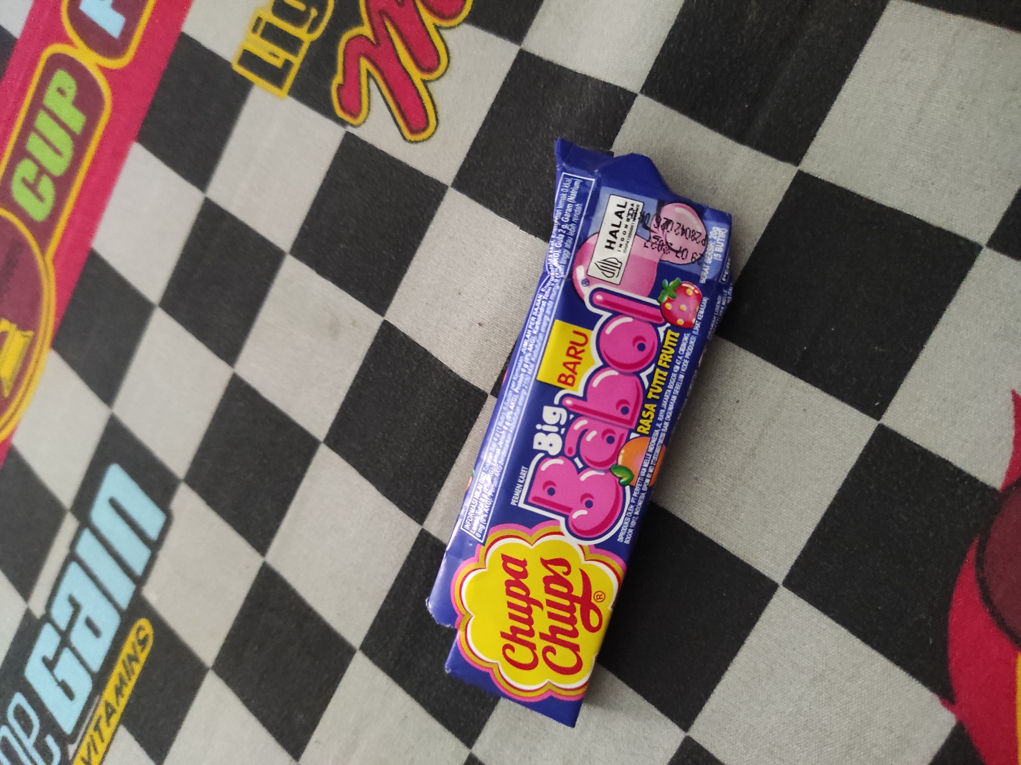 The image shows a Chupa Chups Big Babol bubble gum wrapper on a black and white checkered surface. The wrapper is blue with pink lettering and features images of strawberries and a tangerine. The text on the wrapper indicates it is a "Big Babol" gum with "Rasa Tutti Frutti" (Tutti Frutti flavor) and is produced in Indonesia. The background appears to be a tablecloth or similar fabric with a checkered pattern, possibly a racing flag design, and some other blurry text elements that are not clearly discernible. The image's lighting is bright, casting some shadows. There is no indication of the city of Sumedang, Indonesia, in the image.