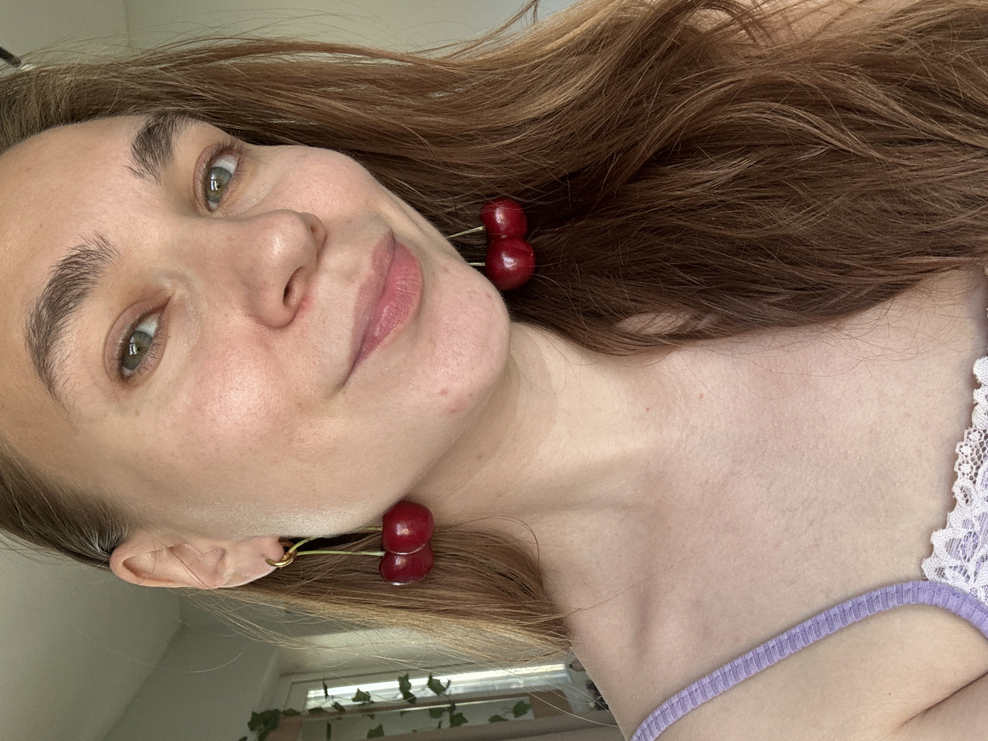 A medium close-up portrait, oriented sideways, features a young woman with light brown hair, captured in what appears to be an indoor setting in Kharnavy, Russia. Her head is tilted significantly, with her gaze directed towards the viewer, accompanied by a subtle, pleasant smile. Her eyes are a distinct green or hazel, and her long hair cascades across the upper part of the frame.
She is wearing unique red, glossy cherry-shaped earrings on both ears; one pair is clearly visible on her left ear, while the other peeks out from behind her hair on her right ear. Her attire includes a light purple or lilac-colored top, possibly a camisole or bralette, featuring delicate white lace trim along the strap on her right shoulder. The lighting is soft and natural, suggesting the photograph was taken during the daytime. Hints of a window and green plant foliage are faintly visible in the soft-focus background, indicating an indoor location with natural light. The woman's expression conveys a sense of calm and approachability. No visible text is present in the image.