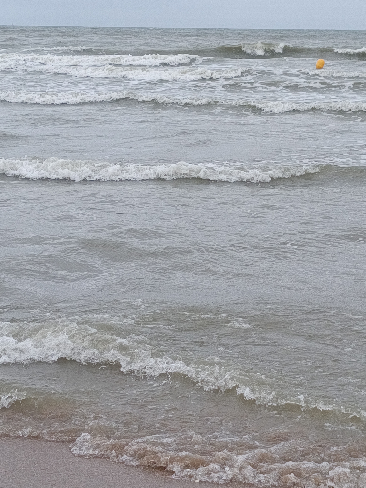 The image depicts a view of the sea at Houlgate, France, with several waves rolling towards the shore. The water is a muted grey, and the sky is overcast, creating a somber atmosphere. The waves are marked by white foam as they crest and break. In the distance, a small, bright yellow buoy is visible floating on the water. The foreground shows a sandy beach with the edge of a wave lapping at it. There are no people or animals visible in the image.