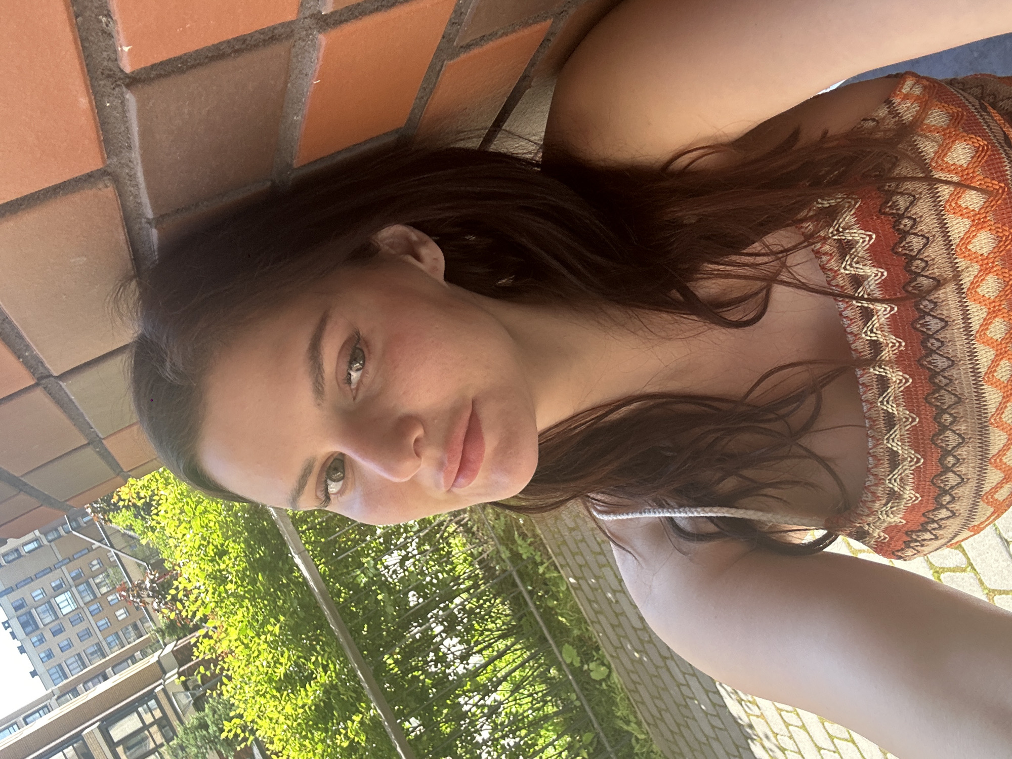 A woman is taking a selfie outdoors. She has long, dark brown hair and is wearing a patterned strapless top. She is leaning against a brick wall, and there are green bushes and a tall building in the background. The ground is paved with cobblestones. The lighting suggests it is daytime.
