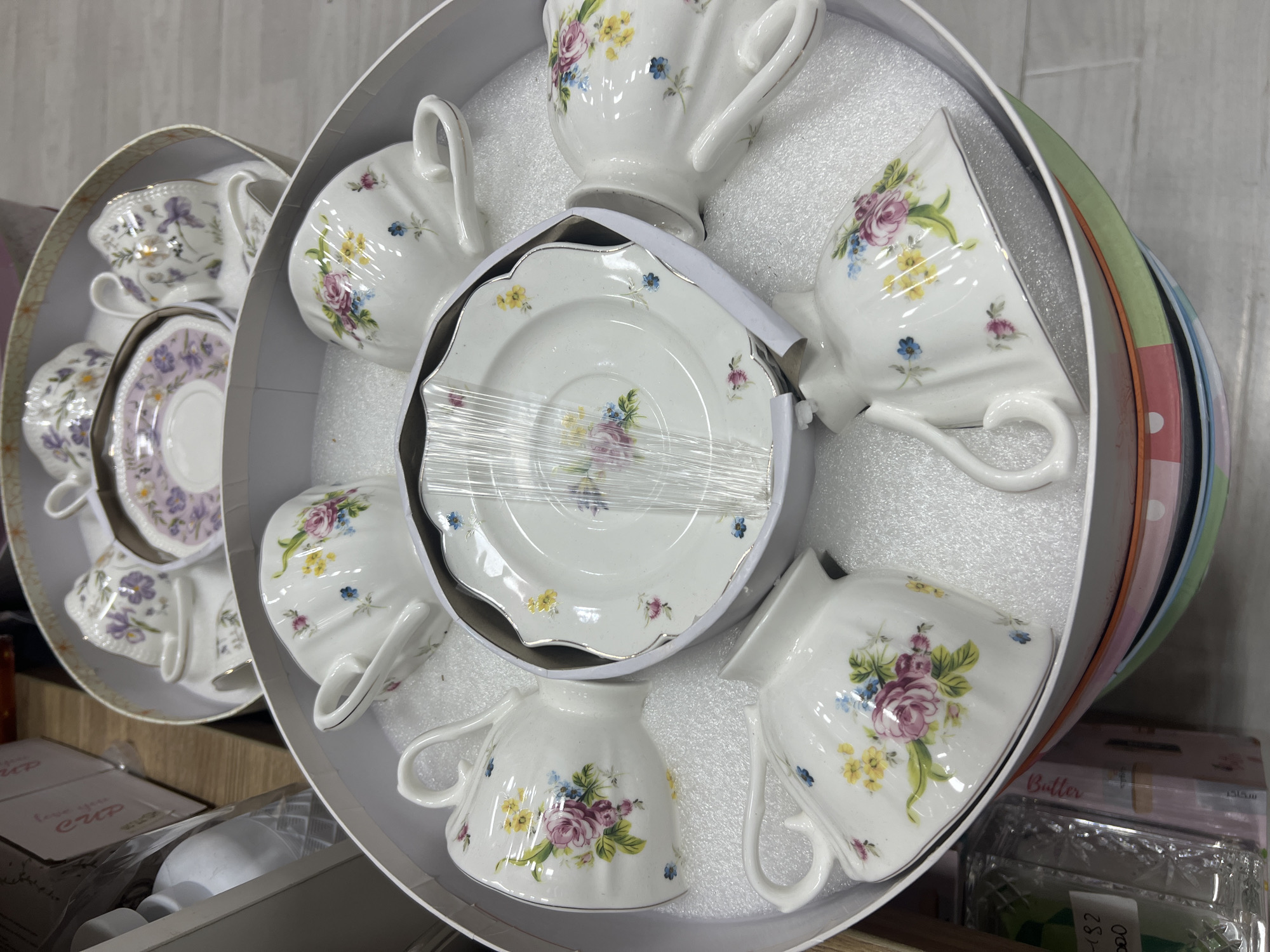 A top-down, close-up, eye-level shot shows a set of fine china teacups and saucers, arranged in a round, white gift box. The teacups and saucers are white, with delicate floral patterns of roses, wildflowers, and greenery. Some of the cups and saucers have a gold trim. The set is displayed in a store, likely in Eshanguzar, Uzbekistan, as indicated by accompanying metadata not present in the image itself. The gift box is placed on top of a stack of colorful, circular plates. In the background, other boxes and packaging materials are visible, suggesting a retail environment. There are no people or animals present in the image.