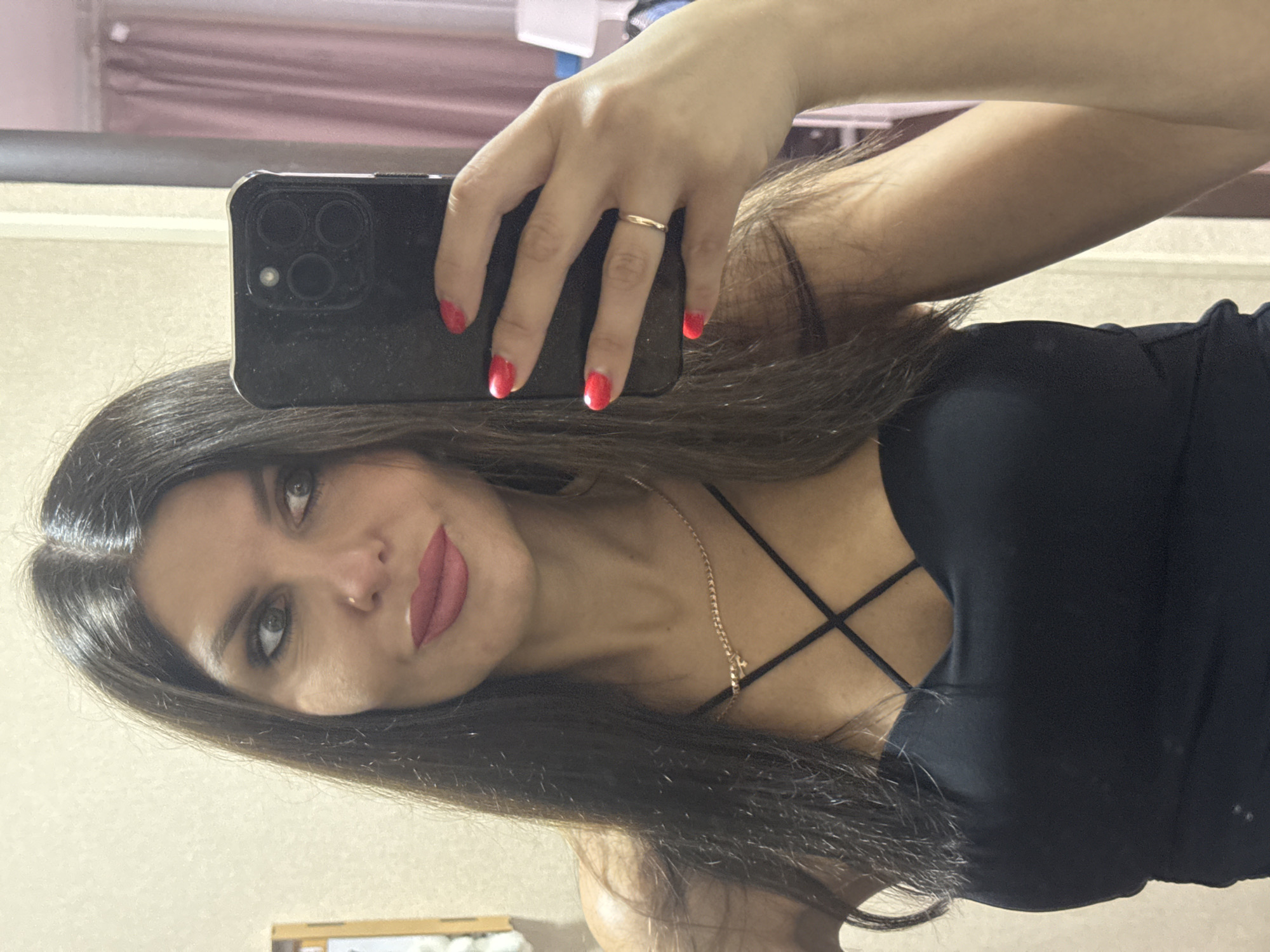 A woman is taking a selfie in a mirror. She has long, dark brown hair, blue eyes, and is wearing a black top with a crisscross neckline. She has red nail polish on her fingers and is wearing a gold ring on her left hand. The background appears to be a room with light-colored walls and some soft pink curtains visible in the mirror's reflection. The woman is holding a black smartphone to take the picture. No specific location in Chaikovsky, Russia is identifiable from the image.