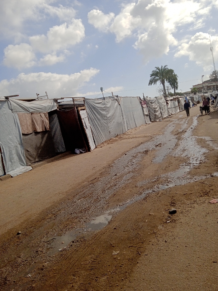 An exterior long shot captures an unpaved dirt road in Khan Yunis, Palestinian Territory, under a sky with scattered white clouds. A prominent row of makeshift shelters, constructed primarily from light grey tarpaulins stretched over frames of wood and possibly metal, lines the left side of the road. Some shelters feature darker fabric panels or exposed wooden components. One shelter exhibits an open entrance with a white object, possibly clothing, at its base.
The reddish-brown dirt road displays multiple muddy patches and standing water puddles, indicating a wet surface. Visible tire tracks are present in the mud. In the mid-ground, on the right side of the road, a person in dark attire walks away from the camera. Further down the road, another individual pushes a two-wheeled cart. Distant figures and additional structures are visible in the background. A tall palm tree stands on the right side, behind the shelters, with multi-story buildings and a communications tower visible further in the distance. Street light fixtures are mounted above some of the shelters.