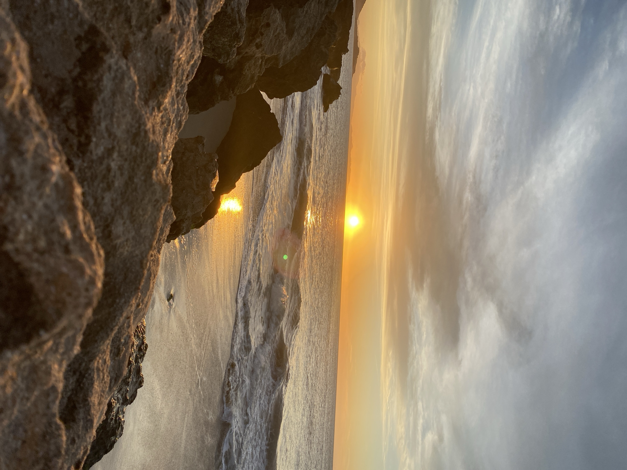 The image shows a sunset view from a rocky area at Playa Caribe in Juan Griego, Venezuela. Large, dark-colored rocks are visible in the left foreground, partially obscuring the view. The ocean stretches out in front, with waves gently lapping the shoreline. The setting sun is positioned in the middle of the horizon, creating a bright glow that reflects on the water's surface. The sky is filled with light clouds tinged with orange and yellow hues from the sunset. The overall scene depicts a tranquil coastal environment at sunset.