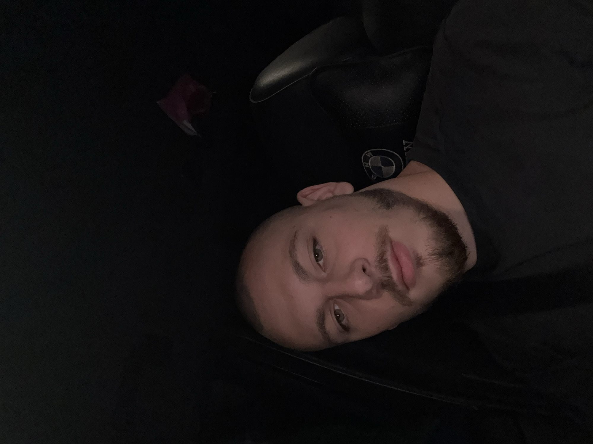 A young man with a buzz cut and a goatee is seated in a car. He is wearing a black t-shirt. The interior of the car is dark, with black leather seats visible. A BMW logo is visible on the headrest of the seat behind him. A pink object is hanging from the car's ceiling. The image appears to be a selfie taken by the man. The location is not discernible from the image.