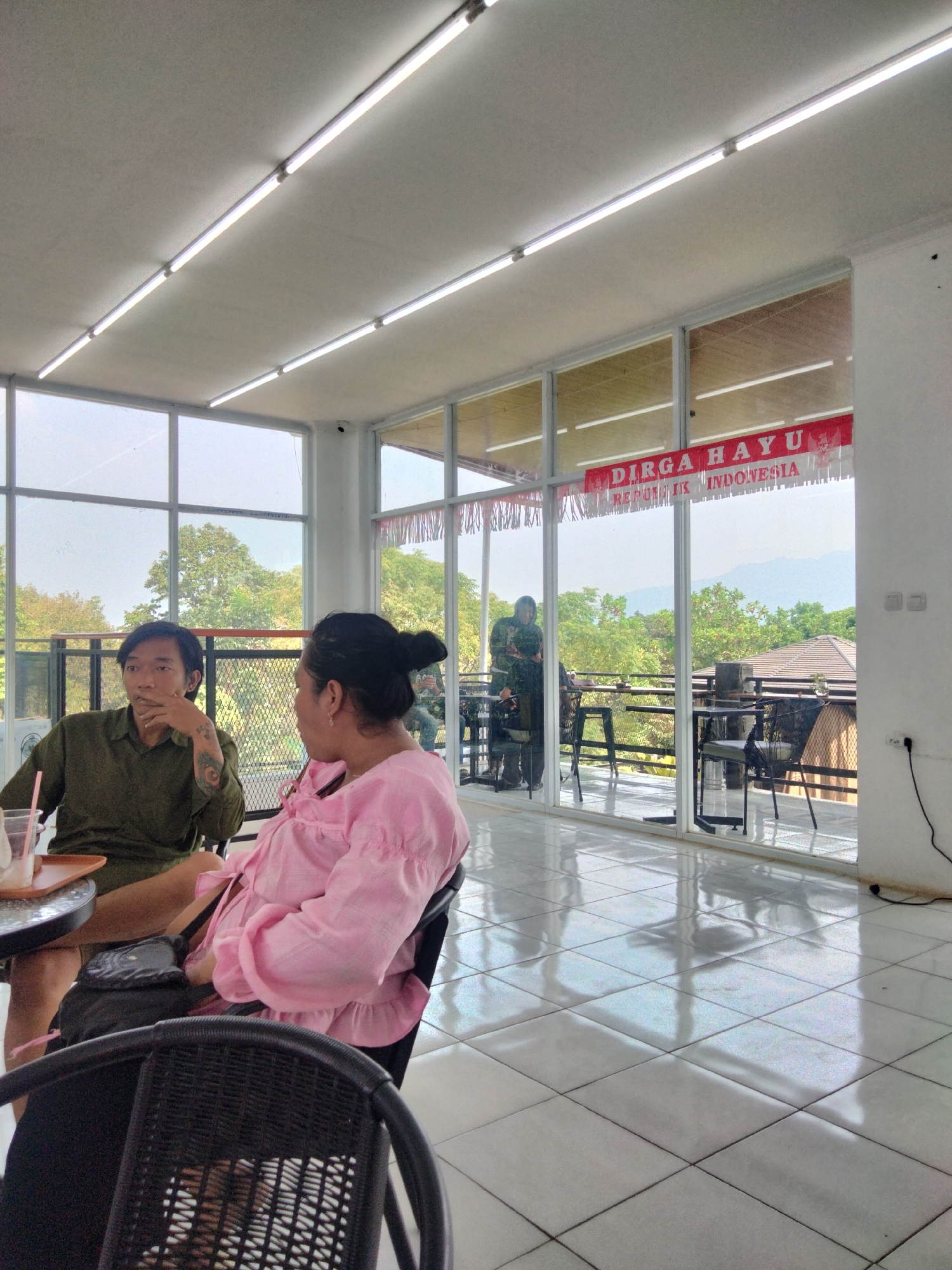 A medium shot shows two individuals, a man and a woman, seated at a table inside a cafe in Tanjungsari, Indonesia. The man, wearing a green shirt, is on the left, looking away from the camera and resting his chin on his hand. He has tattoos on his left arm. The woman, in a pink blouse with ruffled sleeves, is on the right, facing away from the camera and engaged in conversation with the man. A black purse is visible on her lap. The cafe has large windows overlooking an outdoor area with trees and mountains in the distance. A red and white banner reading "DIRGA HAYU REPUBLIK INDONESIA" hangs across the top of the windows. The interior is brightly lit by fluorescent lights on the ceiling and the white tiled floor is highly reflective. The atmosphere appears relaxed, with the individuals seemingly in conversation.