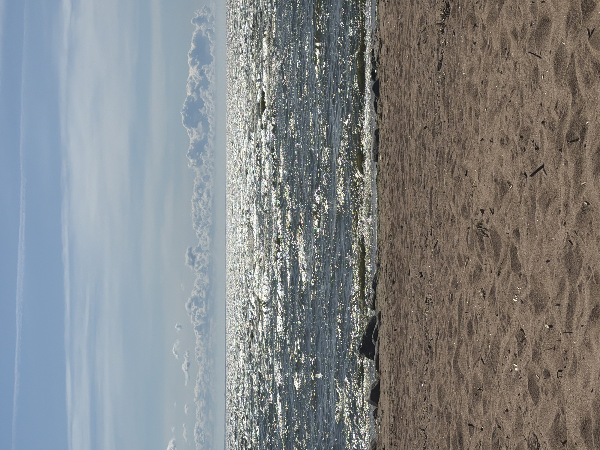 The image depicts a sandy beach in Repino, Russia, leading to the Gulf of Finland. The foreground is dominated by textured brown sand, with scattered small twigs and debris. Along the waterline, there are a few dark rocks. The water is dark blue and choppy, with sunlight glinting off the surface, creating a shimmering effect. In the distance, a line of puffy white clouds is visible against a bright blue sky, with thin, wispy contrails streaking across the upper portion of the sky. There are no people or animals visible in the scene.