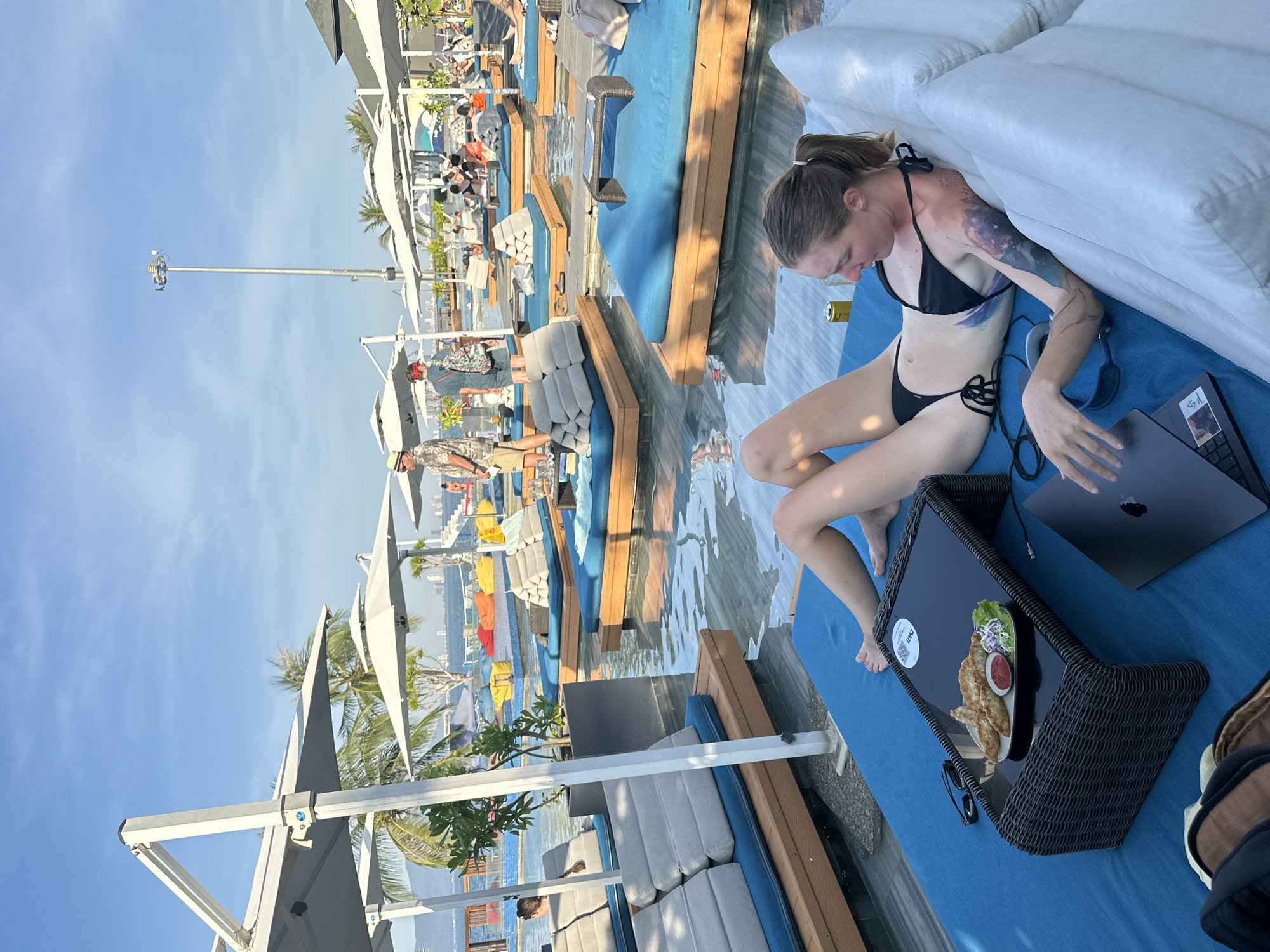 The image shows a woman in a black bikini sitting on a blue lounge chair at a resort in Da Nang, Vietnam. She is looking down at a laptop on a wicker tray in front of her, which also holds a plate of food and a can. The woman has tattoos on her arm. The scene is set outdoors with clear blue skies and bright sunlight, suggesting it is daytime. In the background, there are other people relaxing on lounge chairs around a pool, and some palm trees are visible. Umbrellas are set up to provide shade. The overall atmosphere appears to be relaxed and leisurely, typical of a vacation setting. The text "DÀ NẴNG" is visible on a sticker on the tray.