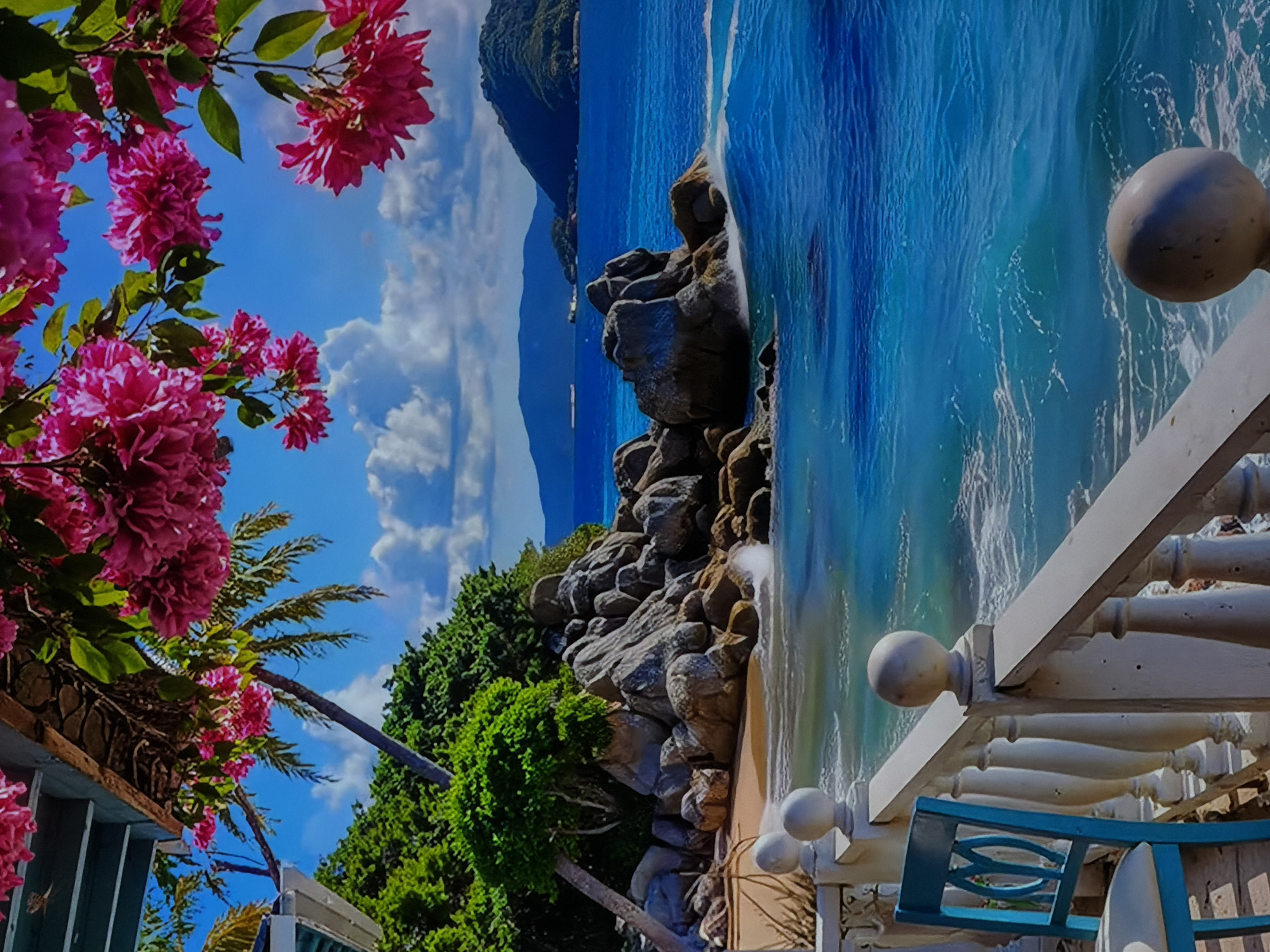 The image, viewed in a landscape orientation, presents a vibrant and picturesque coastal scene, likely from a balcony or elevated viewpoint. In the foreground, rich clusters of bright pink and fuchsia flowers with green leaves adorn the upper left and left edges, framing the view. A white balcony railing with decorative balusters and spherical finials extends from the bottom right, with a portion of a blue outdoor chair visible below it.
The midground reveals a rugged coastline composed of large, grey rocks, interspersed with lush green tropical vegetation, including palm trees. A small stretch of sandy beach is visible along the base of the rocks. The dominant feature is the expansive, clear turquoise ocean, with gentle waves breaking near the shore, creating white foam. In the distant background, rolling hills or mountains are visible under a bright blue sky dotted with scattered white clouds. A small, dark object, possibly a boat, can be discerned in the distant water.
The scene suggests a sunny daytime, with clear weather and bright natural light. The overall atmosphere evokes a tropical or subtropical paradise, characterized by vibrant colors and natural beauty. There are no people or specific activities depicted, and no visible text. The visual cues in the image strongly suggest a tropical coastal region with abundant foliage and clear waters. The provided location context is Sofiivska Borshchahivka, Ukraine.