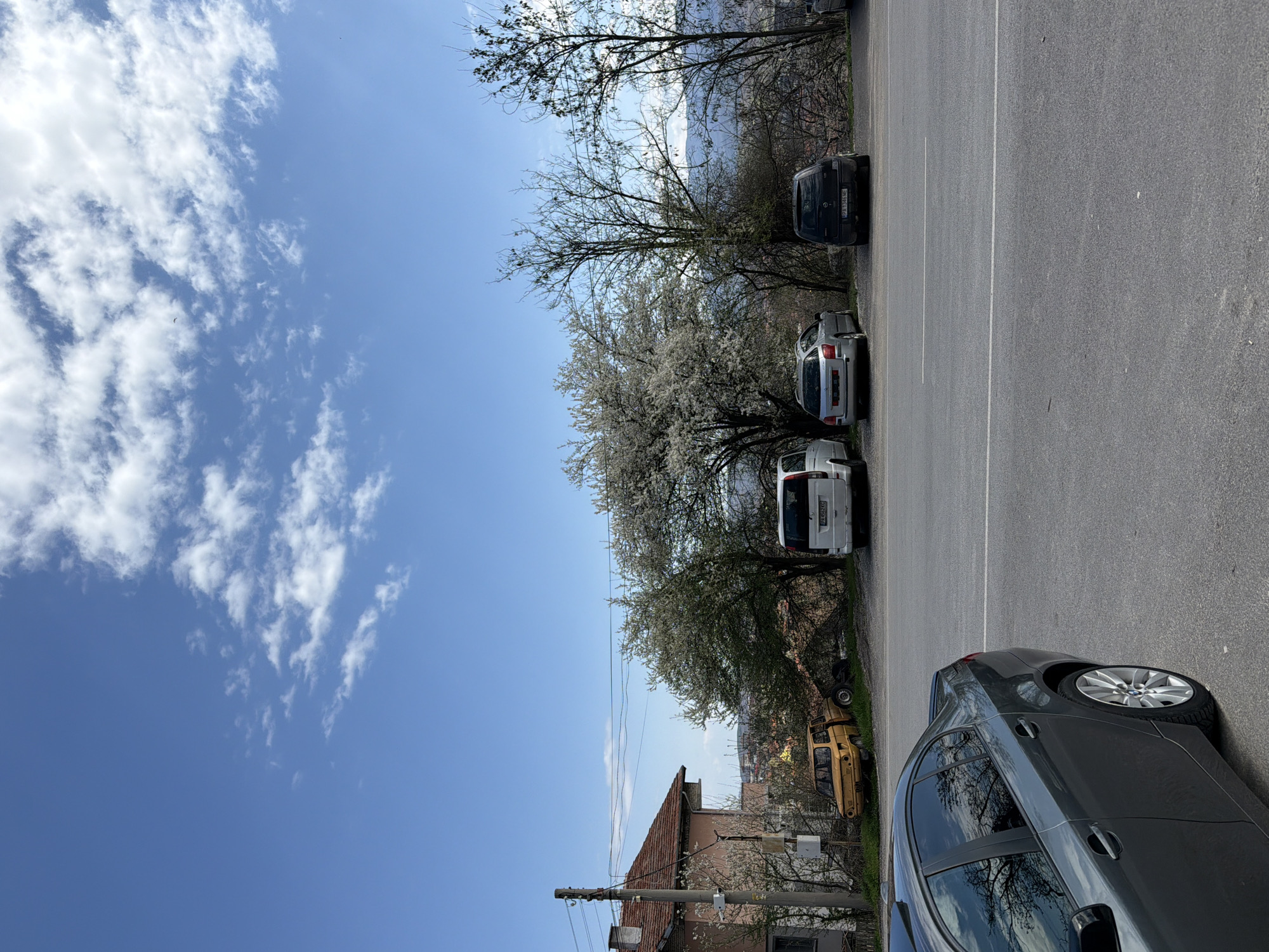 The image shows a street scene in Sveti Konstantin, Bulgaria. The view includes a portion of a building and utility pole at the bottom. Parked parallel along the edge of the road are five cars, with a grey sedan in the immediate foreground. Behind the grey car are two white vans, a silver car, and a black van. Trees are situated between the parked cars and a glimpse of buildings and scenery in the distance. The sky, which occupies a significant portion of the image, is blue with scattered white clouds. The road is asphalt with painted white lines. The photograph appears to have been taken during the day, under natural lighting conditions.