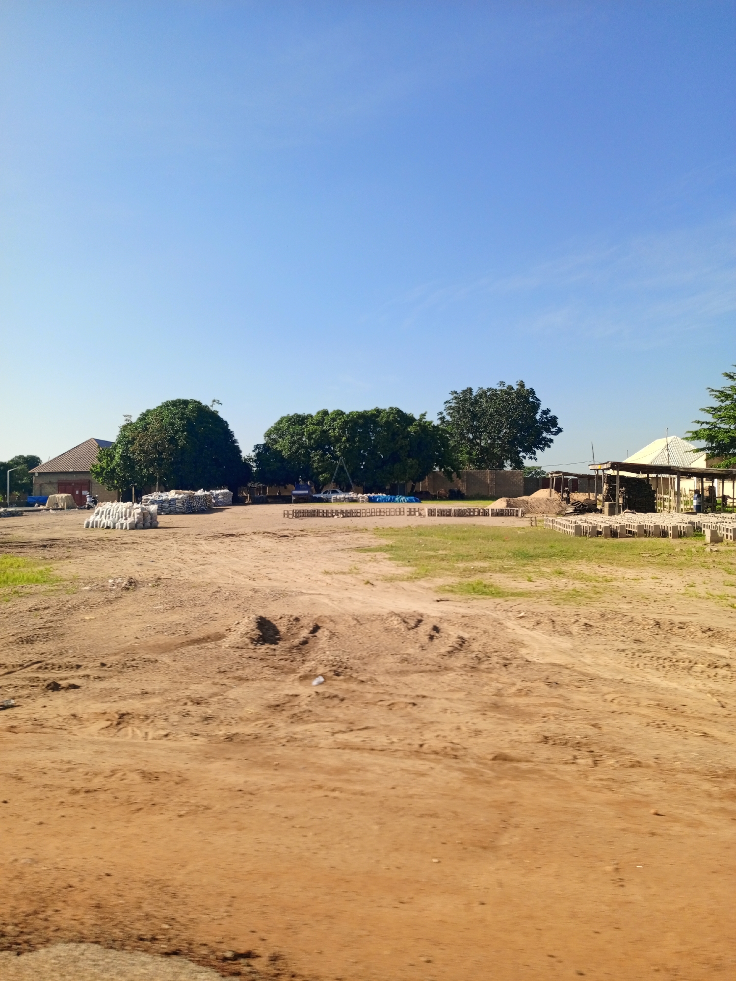 The image depicts an outdoor scene in Toro, Nigeria, likely a construction or material storage area. The foreground is dominated by a sandy, uneven ground with tire tracks visible. In the midground, several stacks of white bags, possibly cement or building materials, are arranged on the ground. Behind these, a building with a pitched brown roof and brick walls stands. Several large, leafy green trees are present throughout the scene, providing shade. Further back, more piles of materials, including what appear to be concrete blocks, are visible, along with a covered structure supported by wooden poles. A white truck and a blue vehicle are parked in the distance, and a person on a motorcycle is seen near the building in the midground. The sky is clear and blue.