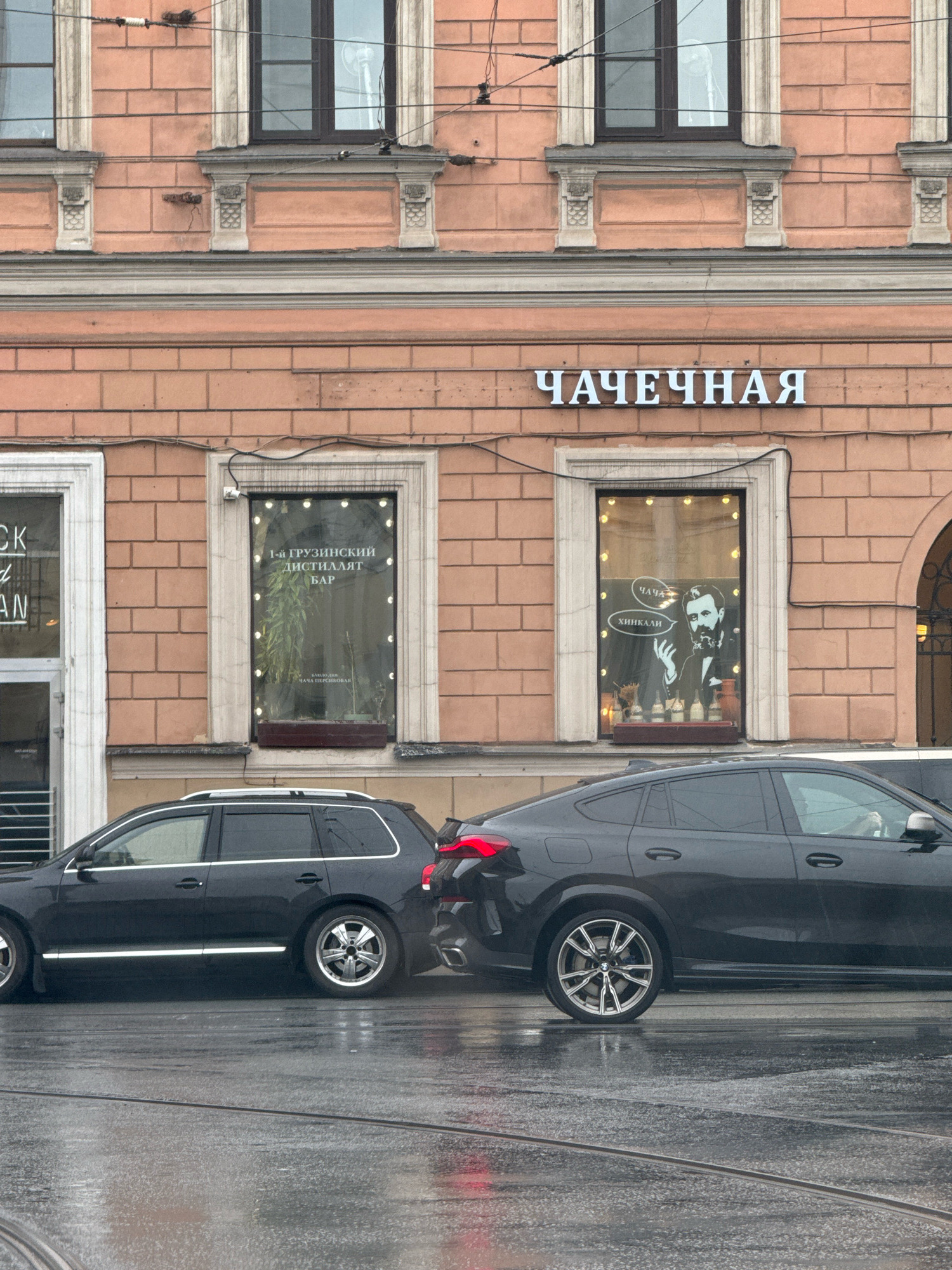 The image captures a street scene in Saint Petersburg, Russia, on a wet, overcast day. In the foreground, two dark-colored cars are parked on a wet street with tram tracks visible. The car on the left is a station wagon, and the car on the right is a coupe with a distinctive angled rear.
Behind the cars is the facade of a building, featuring a light salmon-colored brick exterior. The building has large windows, some of which display signage and decor. One window has the Cyrillic text "ЧАЧЕЧНАЯ" illuminated above it, indicating a tea house or a place that serves tea. Another window displays text that appears to read "1-й ГРУЗИНСКИЙ ДИСТИЛЛЯТ БАР" and "ЧАЧА ПЕРСНИКОВАЯ," suggesting a Georgian establishment, possibly a bar specializing in chacha, a Georgian spirit. The same window also features a silhouette of a man with speech bubbles containing "ЧАЧА" and "ХИНКАЛИ," further reinforcing the Georgian theme.
The building's architecture includes decorative stone elements around the windows and a classical cornice. The overall impression is that of a commercial street in a historic urban area.