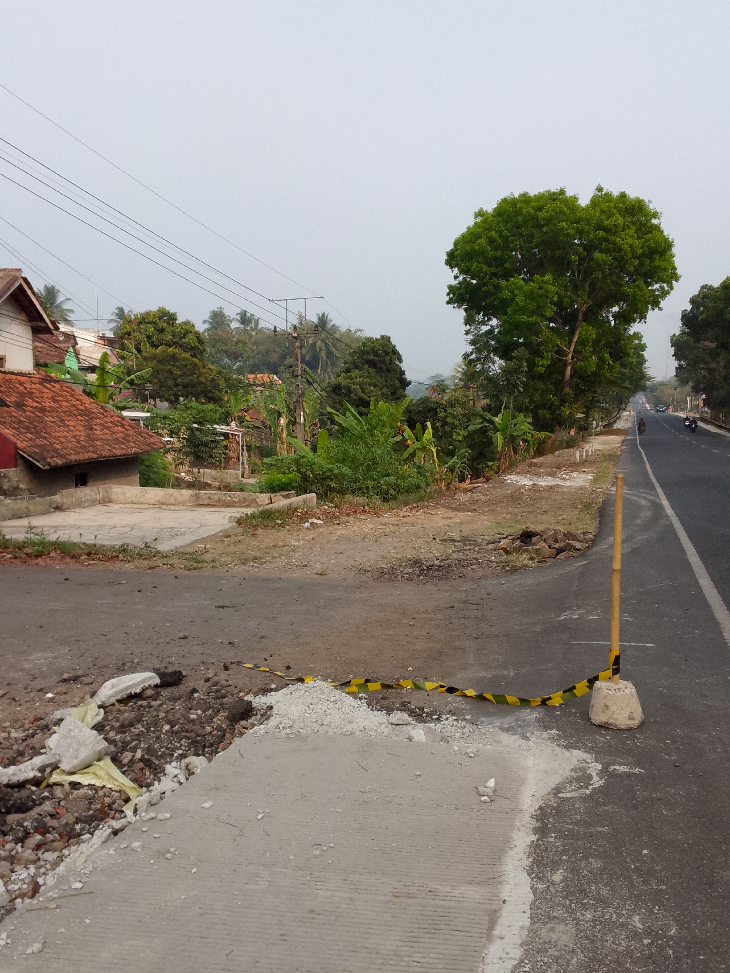 A rural roadside scene in Rajamandala Kulon, Indonesia, shows a section of asphalt road undergoing repair. In the foreground, a new concrete surface is being laid, with piles of rubble and sand nearby. A temporary barrier of yellow and black caution tape, attached to a bamboo pole embedded in a concrete base, demarcates the construction area. Beyond the repair site, the road continues, with a few motorcycles visible in the distance. The roadside is lined with lush green vegetation, including banana trees and other tropical plants. Several houses with reddish-brown tiled roofs are scattered amongst the trees, suggesting a residential area adjacent to the road. Overhead, power lines stretch across the frame against a hazy sky. The overall impression is one of ongoing infrastructure development in a tranquil, semi-rural setting.