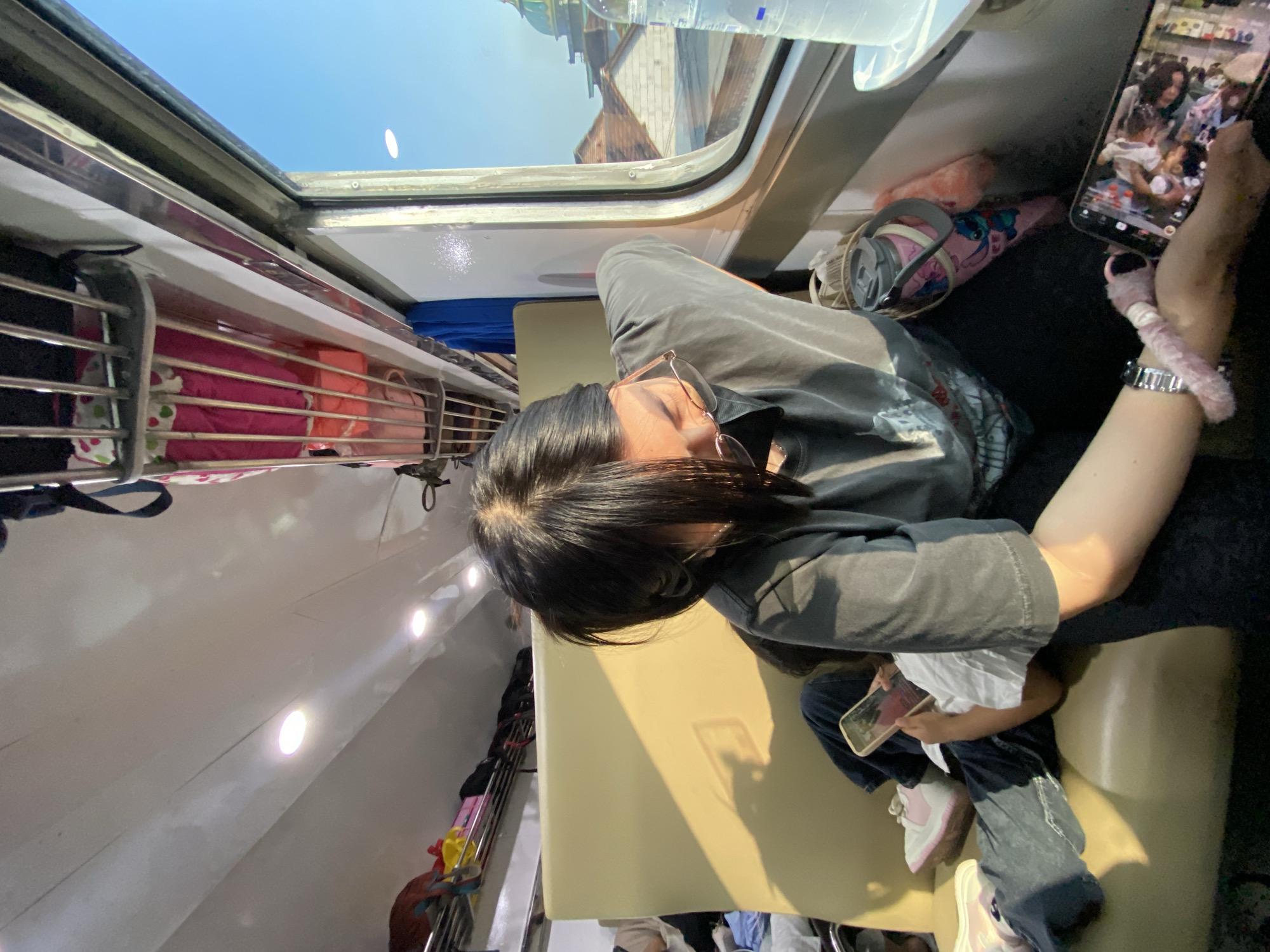 Inside a train, a person is lying down on a seat looking at a smartphone. Another person sits beside them. Visible items include water bottles, a water container, and luggage racks with luggage. Outside the train window, the tops of buildings are visible, suggesting an urban environment in Raci, Indonesia.