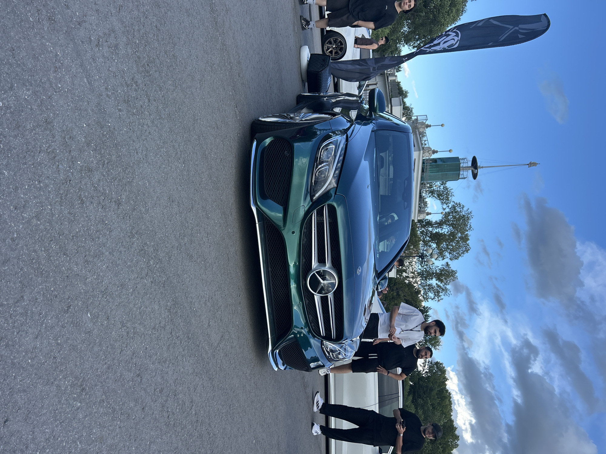 A dark green Mercedes-Benz C-Class sedan, modified with a lowered suspension and custom wheels, is parked on an asphalt surface. The car is positioned at an angle, facing slightly towards the right of the frame. The front grille features the prominent Mercedes-Benz logo.
Several people are standing around the car. To the left, a young man in a black t-shirt and shorts stands near a tall, dark flag with white lettering. Further back, a white car with custom wheels is visible.
In the background, a tall, green, multi-story structure with a pointed top and a weather vane stands against a blue sky with scattered clouds. Trees and some white buildings are also present in the background.
The overall scene suggests an outdoor car event or gathering. The presence of the distinctive architectural structure in the background with a tall tower and flagpole, along with the general ambiance, is consistent with locations like the Amur Bay embankment or a waterfront park in Khabarovsk, Russia.