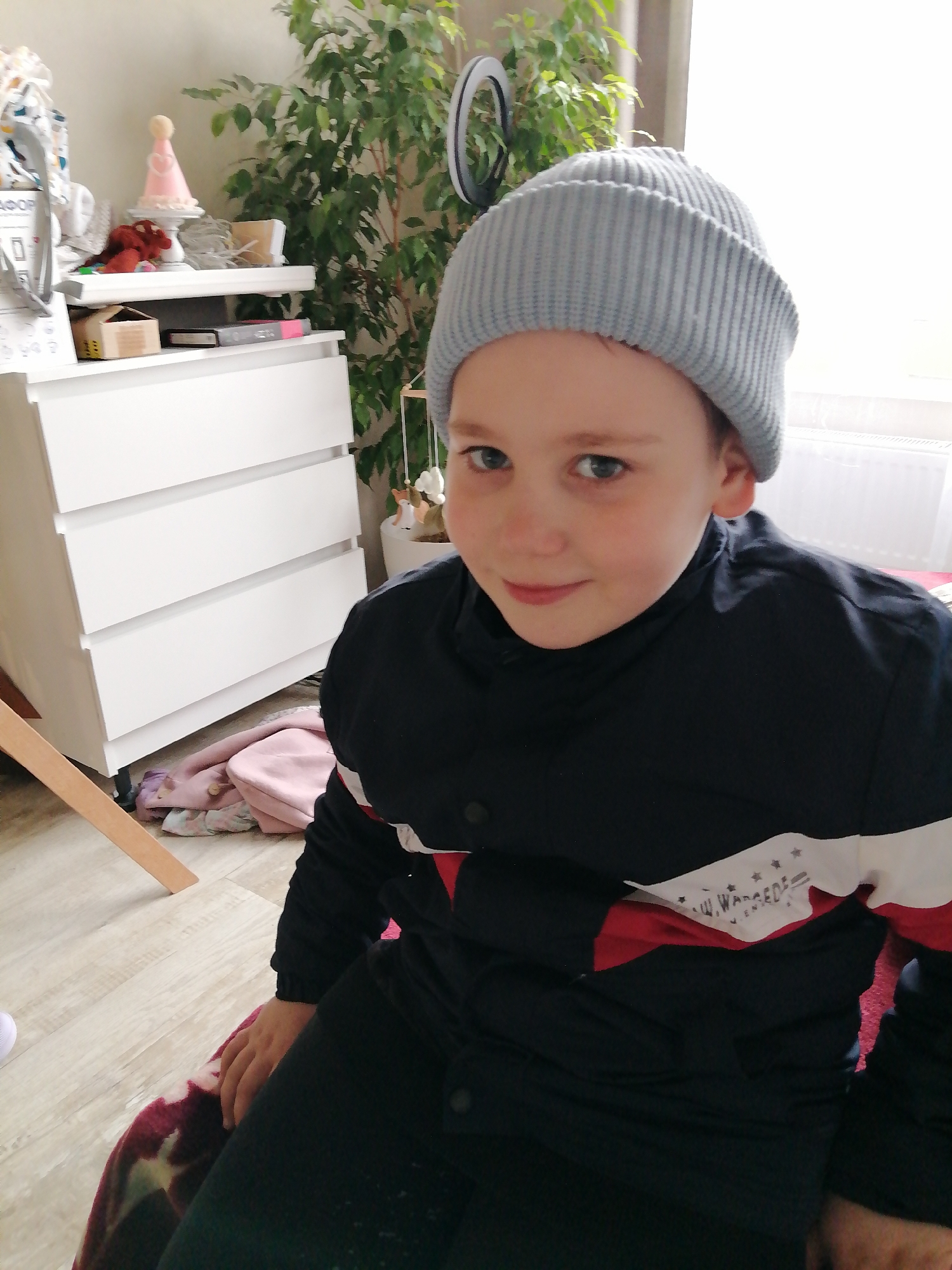 A young boy is seated indoors in Unknown, Unknown. The boy is wearing a gray ribbed beanie and a black jacket with a white and red stripe across the chest. Behind him is a white dresser with three drawers, topped with various objects including a pink cone-shaped hat. A large green plant stands behind the dresser. A portion of a wooden object, possibly a chair leg, is visible on the left side of the frame. The floor is light-colored wood.