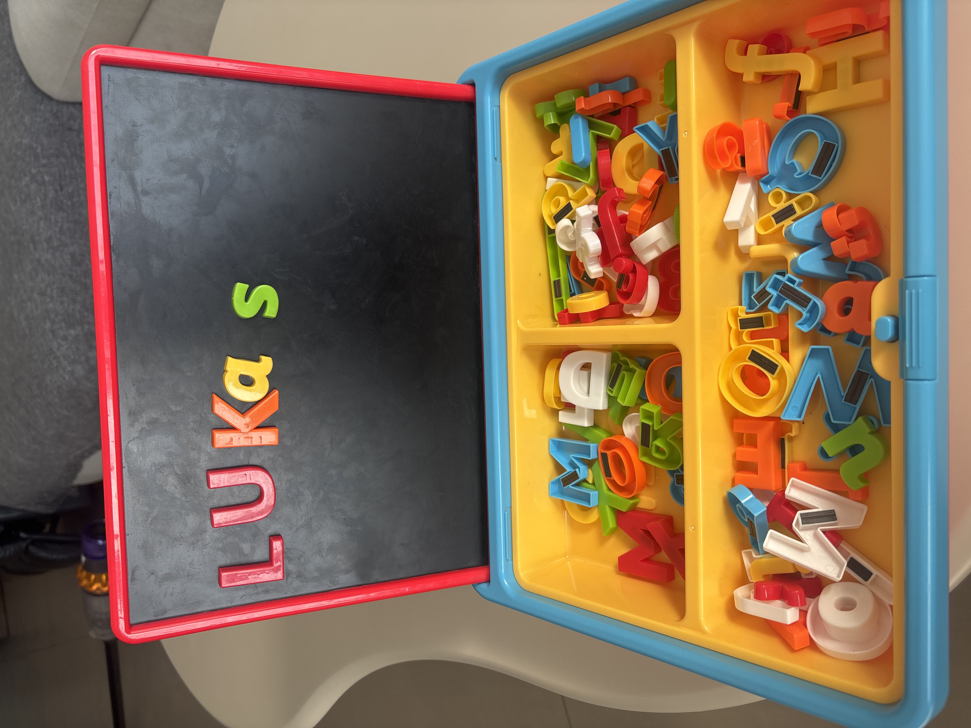 This is a top-down, eye-level shot of a toy alphabet and number set. The set features a black magnetic board with a red frame, propped open to reveal a yellow plastic storage tray. The tray is divided into two sections, both filled with colorful magnetic letters and numbers. The letters on the board spell out "LUKAS". The setting appears to be indoors, likely a home environment, with a blurred background of a grey couch and a white chair. There are no people or animals visible in the image. The image has been taken at an angle that showcases the toy set. The specific location in Dubai, United Arab Emirates cannot be determined from the image alone.