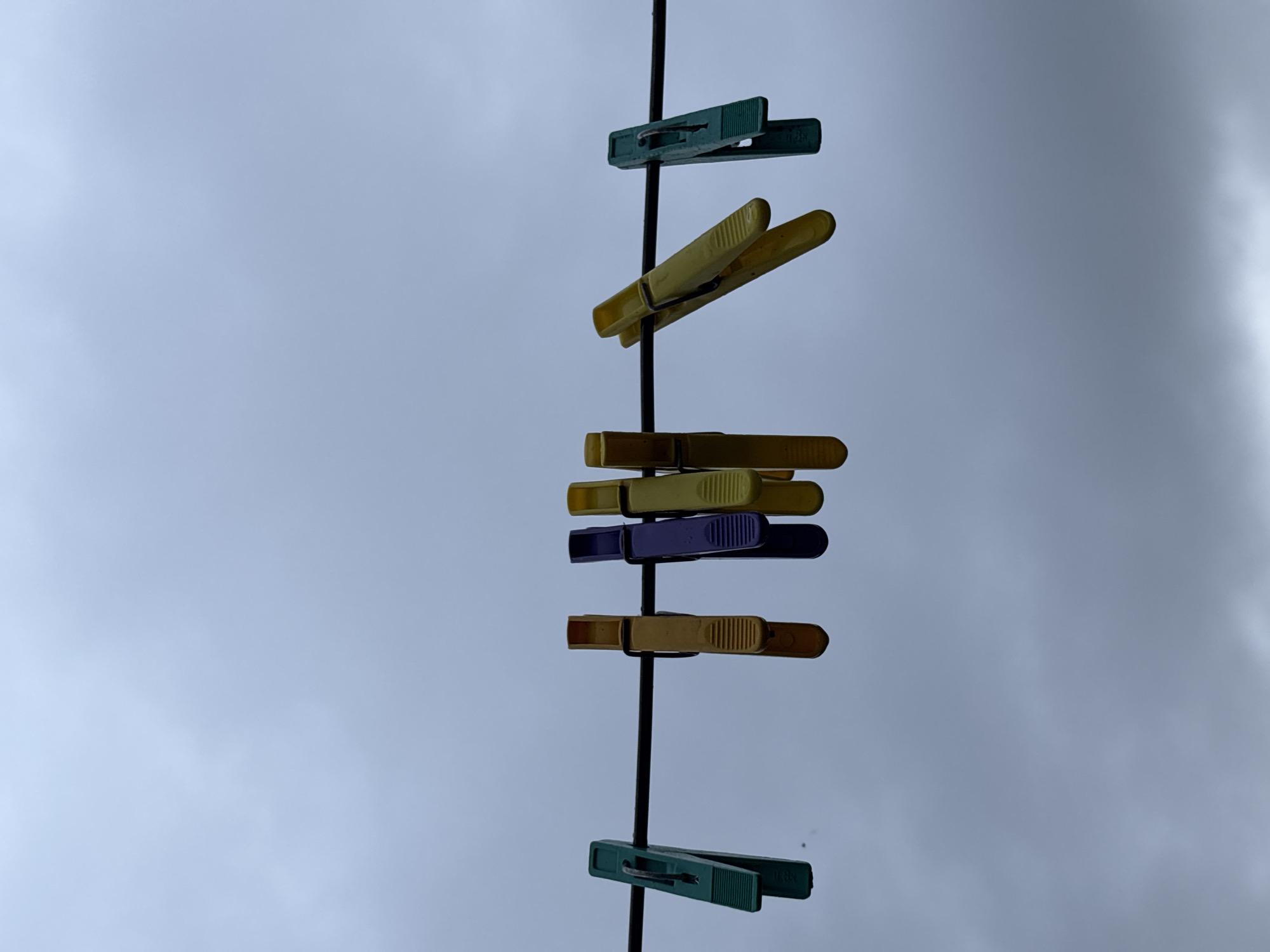 The image shows a close-up, low-angle view of six colorful plastic clothespins attached to a thin, black clothesline. The clothespins are arranged from left to right: one green, two yellow, one purple, one yellow, and one green. The background is a cloudy, overcast sky. The scene is set in Gonja, Russia. There are no people or actions depicted.