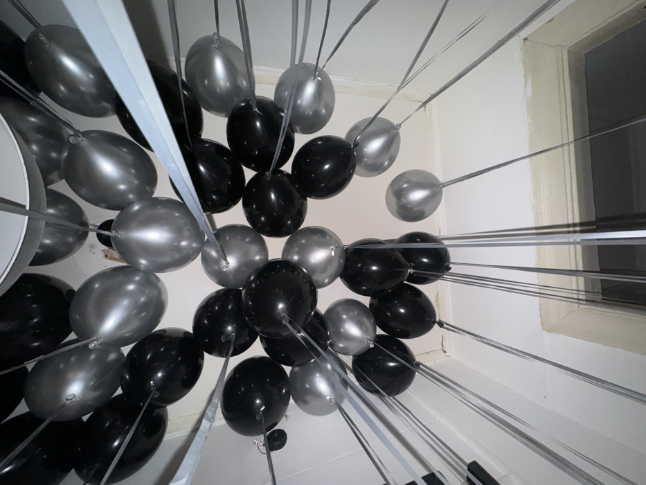 The image displays an indoor scene in Moscow, Russia, primarily focused on a dense cluster of approximately 50-60 balloons suspended near a white ceiling. The balloons are uniformly distributed between opaque black and metallic silver-grey hues. Each balloon is affixed with a thin, grey ribbon extending downwards into the lower frame. A circular ceiling fixture, possibly a light or vent, is partially visible and obscured by balloons in the upper central portion of the ceiling. The surrounding walls are white. A segment of a white-trimmed interior doorway or window frame is discernible in the lower right quadrant, revealing a darker area beyond it. The photograph is captured from a low-angle perspective, directed upwards towards the ceiling.