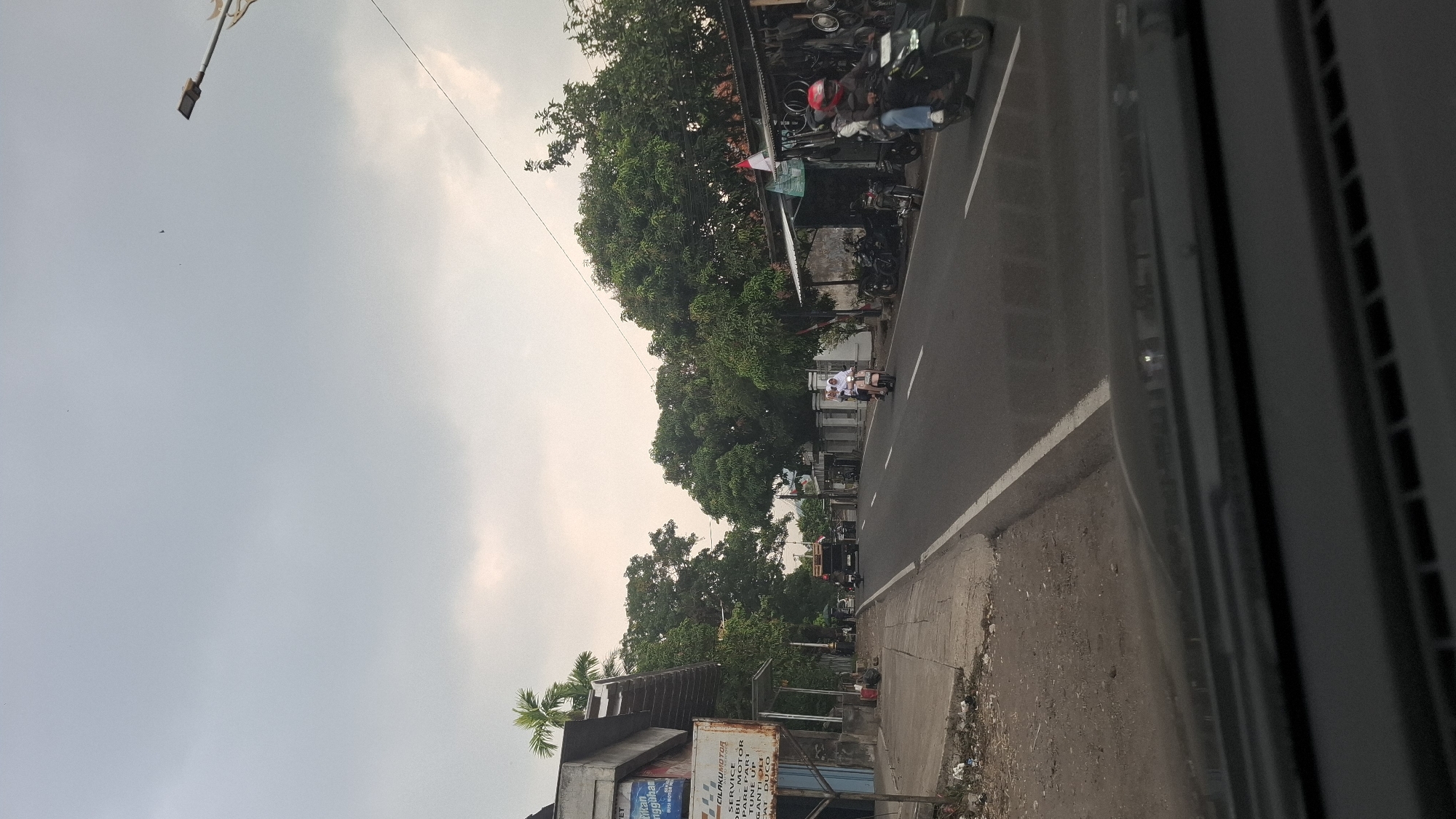 The image captures a street scene in Sukasari, Indonesia, under an overcast sky. The road is paved, with a white line marking the lane division. On the left side of the road, a sidewalk is visible, bordered by a dirt embankment with scattered debris. Several buildings line the street, including a commercial establishment with signage partially obscured. On the right side, trees with dense foliage create a natural canopy over a row of smaller structures, possibly shops or stalls, where motorcycles are parked. Several individuals are present, primarily on motorcycles. In the foreground, two people are riding a scooter, and further down the road, another motorcycle with two riders is approaching. A truck is also visible on the road. The overall impression is one of a typical day in a bustling urban or semi-urban environment.