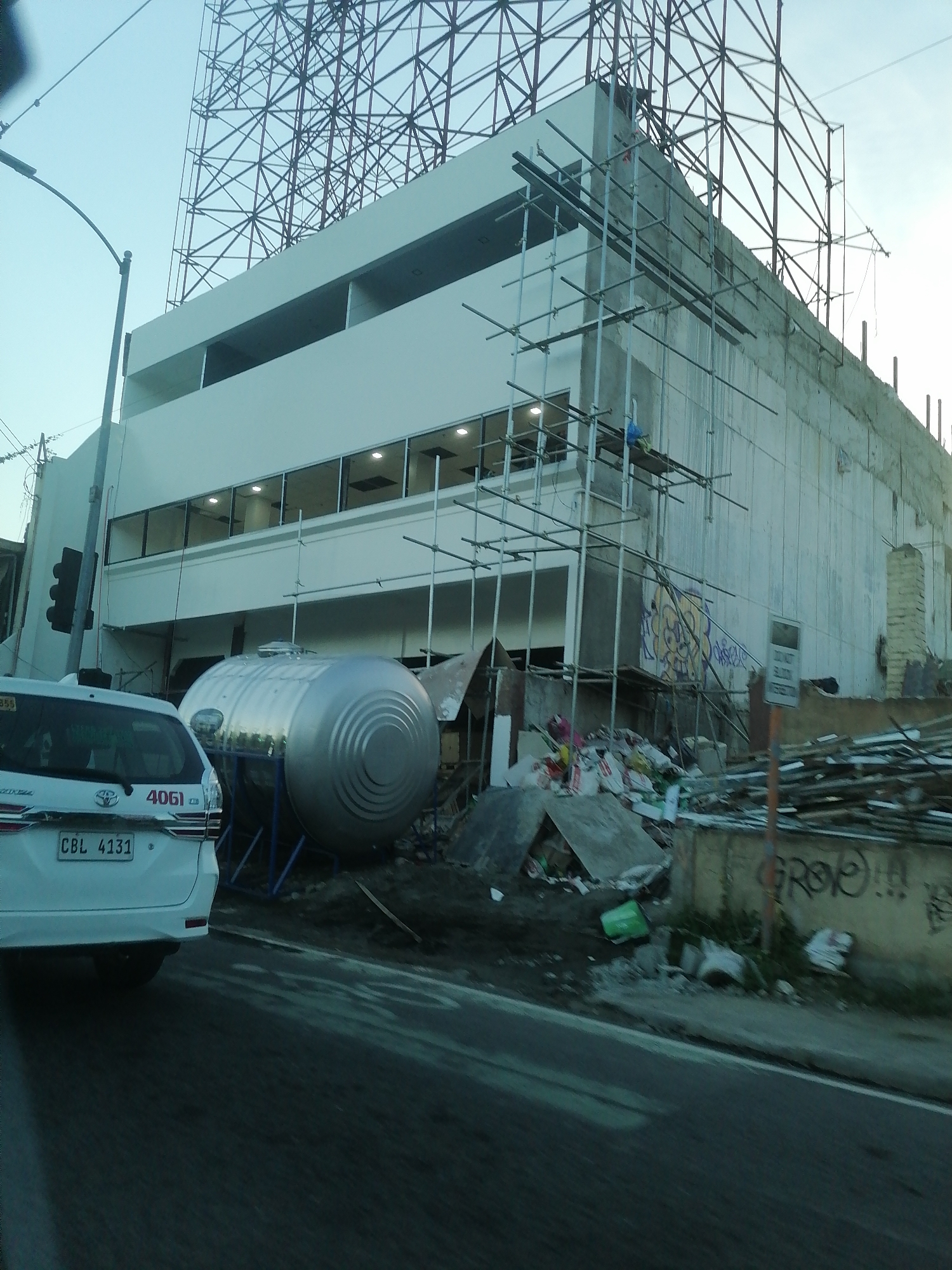 This image captures a daytime scene of an urban commercial area under construction or renovation in Davao City, Philippines. The central focus is a multi-story building featuring a modern aesthetic on its lower levels with a clean white facade and horizontal rows of rectangular windows, some of which are illuminated. The upper right portion of the building appears to be in an earlier stage of construction, displaying exposed concrete and extensive scaffolding. Above the building, a large, intricate metal framework, likely for a billboard or signage, dominates the skyline against a clear blue sky.
In the lower left foreground, the rear of a white Toyota Innova (plate number CBL 4131, with "4061" and "P.U.V." stickers) is partially visible, facing away from the viewer. To its left, a black street lamp and a traffic light displaying red are present. In the mid-ground, positioned near the building, a large, reflective silver horizontal tank, possibly for water storage, rests on a blue metal support structure. The immediate foreground on the right side is cluttered with piles of construction debris, scrap wood, and miscellaneous refuse. This refuse extends along a concrete retaining wall, which is adorned with black and yellow graffiti, including the legible tag "GROO!!!". A partially obscured white sign, indicating a prohibition against littering, stands amidst the debris. The immediate foreground consists of a dark asphalt road with subtle lighter markings.