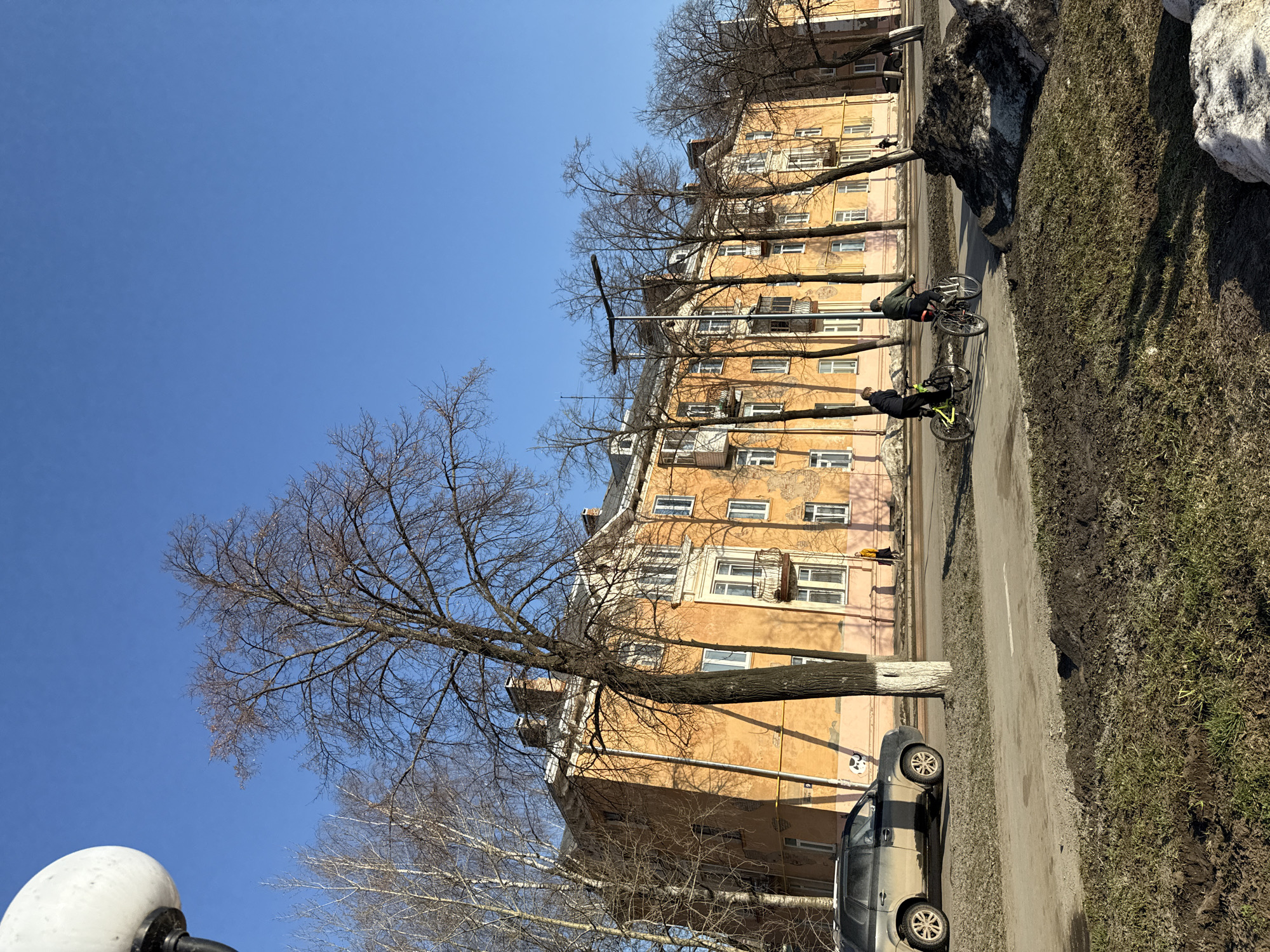 The image shows a street scene in Votkinsk, Russia. A yellow apartment building with white window frames is situated along the right side of the frame. Leafless trees line the street in front of the building. Two people are riding bicycles on the paved road in front of the building. A car is parked along the side of the road near the building. Patches of grass and melting snow are on the right side of the road. The sky is clear and blue. A street lamp is visible in the bottom left corner of the image.