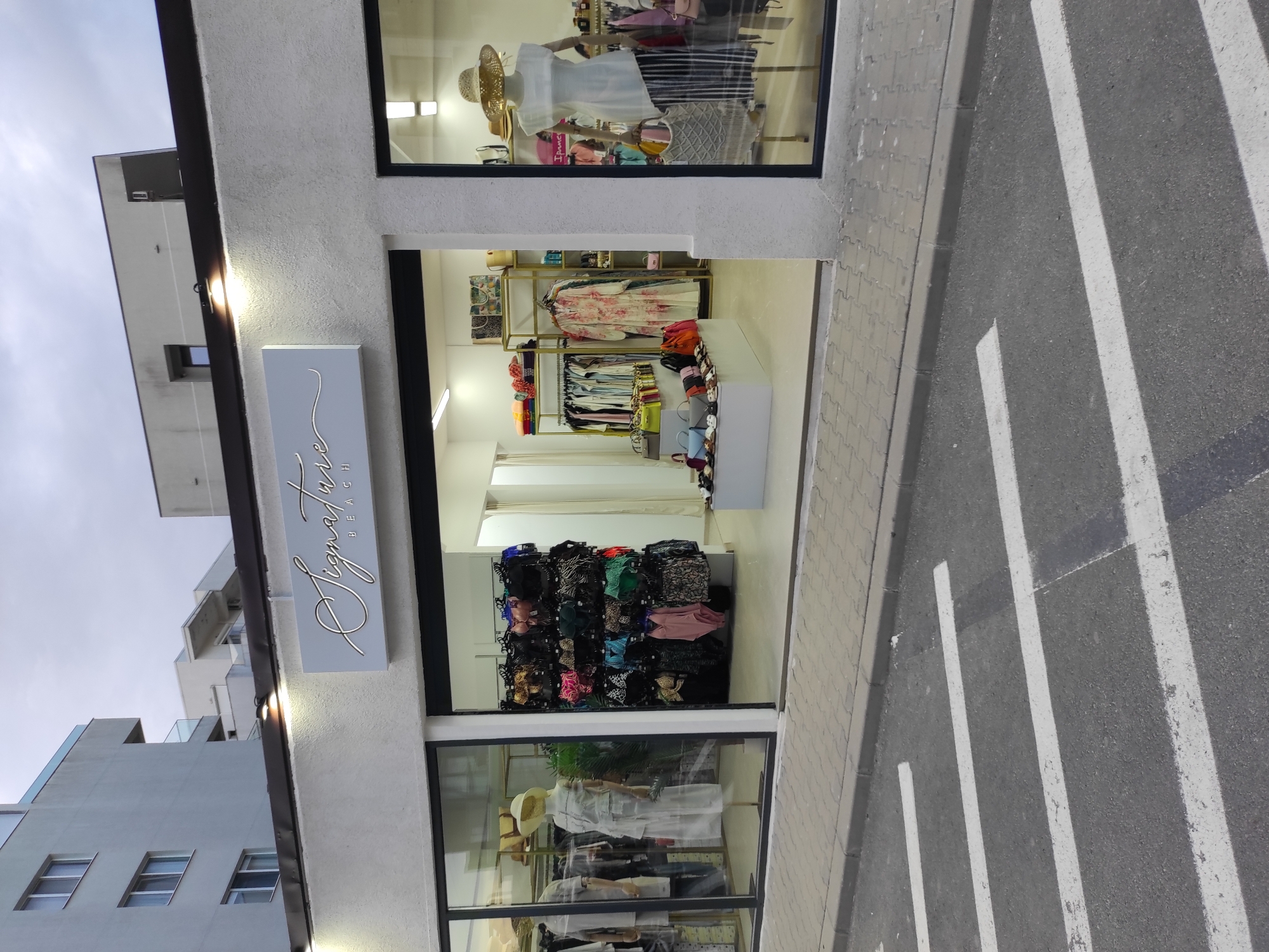 The image shows a street-level view of a boutique store with the signage "Signature BEACH" above the entrance. The store has large glass windows displaying merchandise, including clothing, handbags, and accessories. Inside, mannequins are dressed in outfits, and racks are filled with various apparel items like swimwear and dresses. The store appears to be located in Năvodari, Romania, as indicated by the street markings and the style of the surrounding buildings. The foreground of the image features a pedestrian crossing with white stripes painted on the asphalt. The sky above is overcast. There are no people visible in the image.