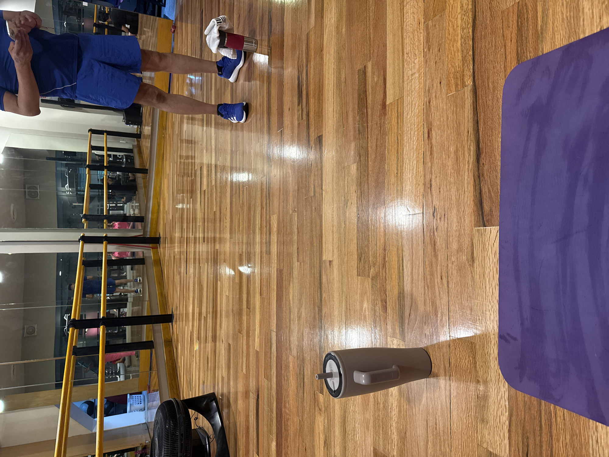 This image captures an indoor fitness studio or gym in Puebla, Mexico, characterized by a polished, reflective wooden floor and mirrored walls. The perspective is from a slightly elevated angle, looking across the facility.
In the mid-ground on the left, a person is partially visible, wearing a blue t-shirt, blue shorts, and blue Adidas sneakers with white soles. They are holding a mobile phone or small device and appear to be looking at its screen, suggesting a break during a workout. Near their feet, a red insulated bottle is visible, partially wrapped in a white towel with a dark, striped logo.
To the right, a purple exercise mat with rounded corners lies flat on the wooden floor. Closer to the foreground, a tall, beige water bottle with a white straw lid rests on the floor. In the bottom-left corner, part of a black standing fan is visible. The mirrored walls reflect various elements of the gym, including yellow and black horizontal bars (possibly for ballet or stretching), other indistinct gym equipment, and reflections of other people in the background. The scene suggests an active, well-lit environment.