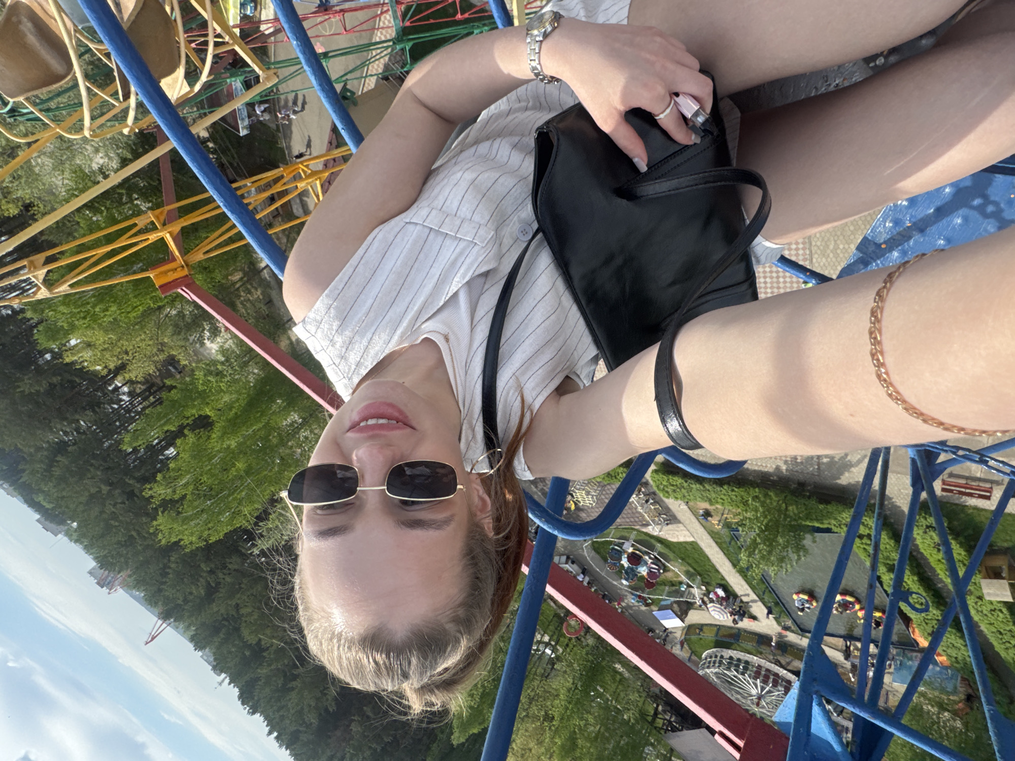 A woman is on a Ferris wheel in Chaikovsky, Russia. She is wearing sunglasses and a striped shirt. She is holding a black purse. The Ferris wheel is painted blue, red, and yellow. There are trees and a park in the background. Other rides and people can be seen in the park below.