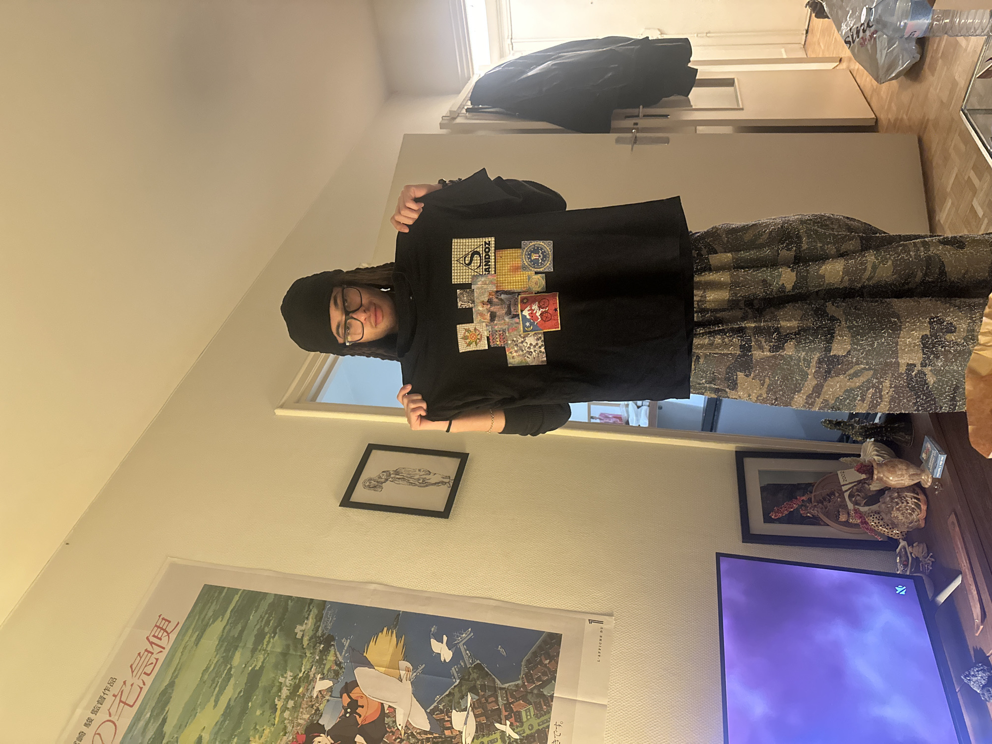 An individual is depicted indoors, holding up a black t-shirt for display within what appears to be an apartment living space. The person, an individual with dreadlocks and dark-rimmed glasses, wears a black beanie and patterned loose-fitting pants, making direct eye contact with the camera. The black t-shirt features a collage of various square and rectangular graphics, including text "SANDOZ," a psychedelic image, and a graphic with a red bicycle and dates "1943 19...".
The surrounding environment includes white walls and wooden parquet flooring. On the left wall, a large framed poster for "Kiki's Delivery Service" displays Japanese characters at the top and the French text "L'AFFICHE DU" at the bottom. A framed drawing of a standing figure hangs on the wall to the right of the poster. In the lower left foreground, a dark flat-screen television displays a purple and blue abstract image, situated on a wooden console adorned with various decorative items such as small statues and vases. An open white door on the right reveals a second room, with a dark jacket hanging on its interior. A glass-top coffee table is partially visible in the lower right, holding a plastic water bottle and a grey plastic bag. The room is illuminated by warm, artificial overhead lighting.
Based on the French text "L'AFFICHE DU" on the poster and typical architectural elements, this scene is located within an apartment in Paris, France.