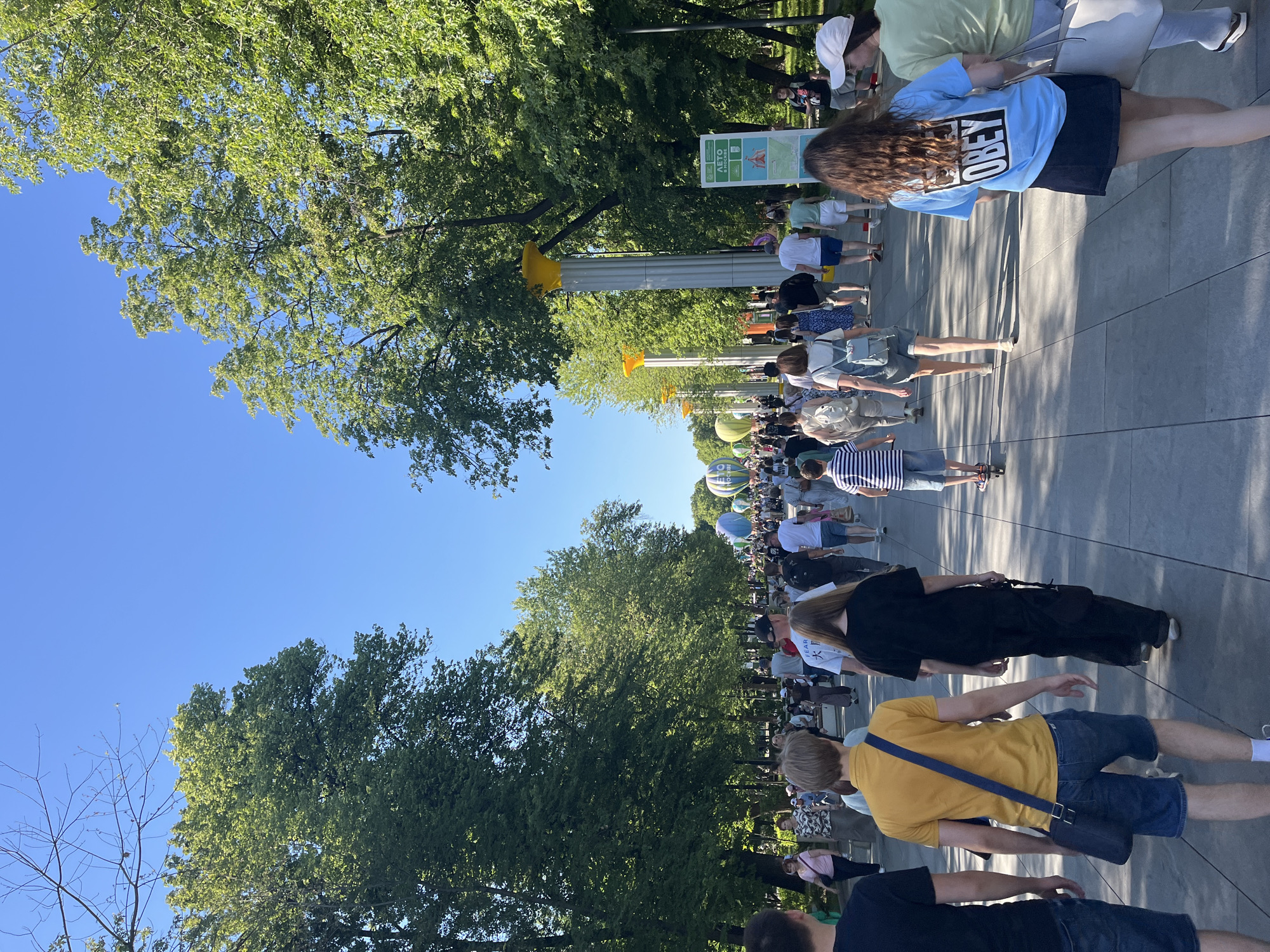 The image depicts a wide, paved pedestrian pathway on a clear, sunny summer day in a park located in Moscow, Russia. The pathway is bordered on both sides by dense, green leafy trees, forming an avenue. On the right side of the path, a series of white columns with distinctive yellow, trumpet-shaped capitals are regularly spaced. A large green vertical sign on the right also displays "ЛЕТО В МОСКВЕ" (SUMMER IN MOSCOW) along with various activity icons and a map.
Numerous individuals of various ages are walking along the pathway, predominantly moving away from the camera. They are dressed in casual summer clothing. In the immediate foreground, from left to right, are the backs of a man in a dark t-shirt and jeans, a man in a yellow polo shirt, blue shorts, and a dark blue messenger bag, and a woman in a black t-shirt and wide-leg black pants. Further back, a woman is visible wearing a light blue t-shirt with the word "OBEY" printed prominently on the back. Other people are seen with backpacks, and one child is on a scooter.
In the mid-ground distance, several large, inflated hot air balloons are visible above the crowd and trees, indicating a special event. One balloon clearly shows the word "МОСКВА" (MOSCOW) written in Cyrillic. The sky is a vibrant, cloudless blue, and bright sunlight casts distinct shadows on the light gray paved surface, which is composed of rectangular tiles. The overall scene suggests a bustling public park during an event or popular leisure day.