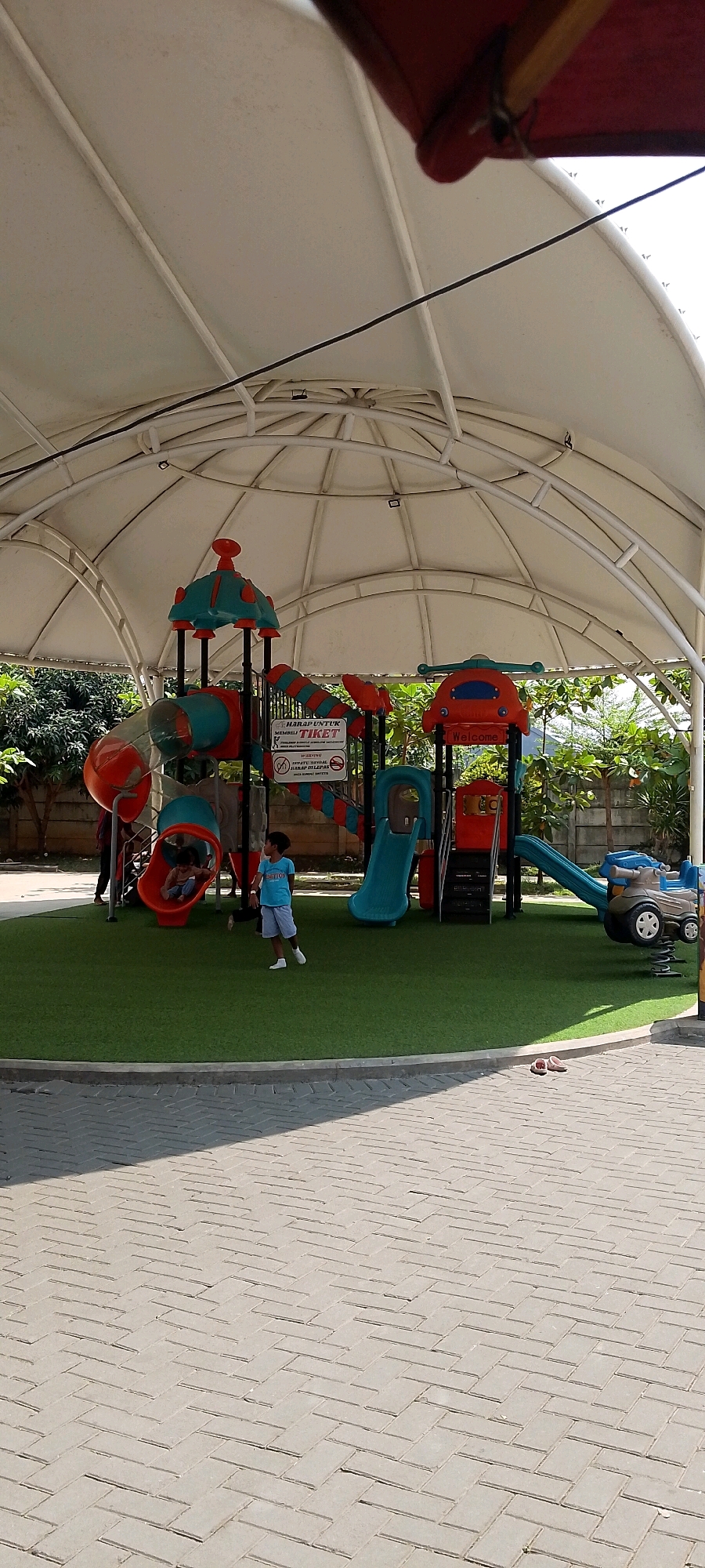 This image depicts an outdoor children's playground under a large, white, domed canopy. The playground features colorful plastic play structures including slides, tubes, and climbing elements in red, blue, and teal. A sign is visible on one of the structures with text in Indonesian, possibly indicating rules or ticket information.
Two children are present. One child, wearing a blue shirt and grey shorts, stands on the green, artificial turf of the playground. Another child, wearing a light brown top, is partially visible inside a red tube slide. An adult, wearing a dark shirt and pants, is visible in the background to the left, near the entrance to one of the play structures.
To the right of the playground, a small toy vehicle resembling a car, mounted on springs, is present. The playground area is bordered by a gray paved walkway made of interlocking brick pavers, which extends to the foreground of the image. The background shows trees and foliage, and a concrete wall. The overall environment appears to be a public space, likely a recreational area or amusement park. The scene is captured in daylight, with shadows cast on the paved area, suggesting it is daytime. The location is identified as Sriamur, Indonesia.