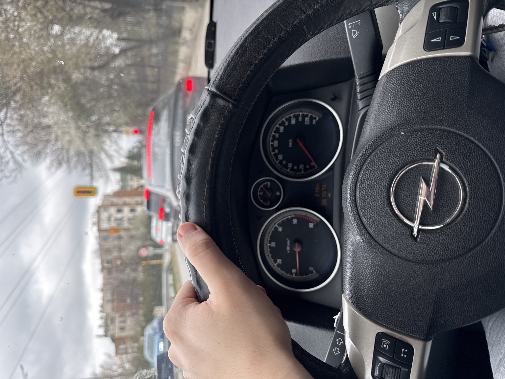 The image shows the interior of an Opel car, with a focus on the steering wheel and dashboard. A hand is partially visible, holding the steering wheel. Through the windshield, traffic and buildings are visible, suggesting an urban setting. Based on the architecture and the road markings visible through the windshield, the location appears to be in Sofia, Bulgaria. The dashboard displays the car's instruments, and some of the screen information is also available.