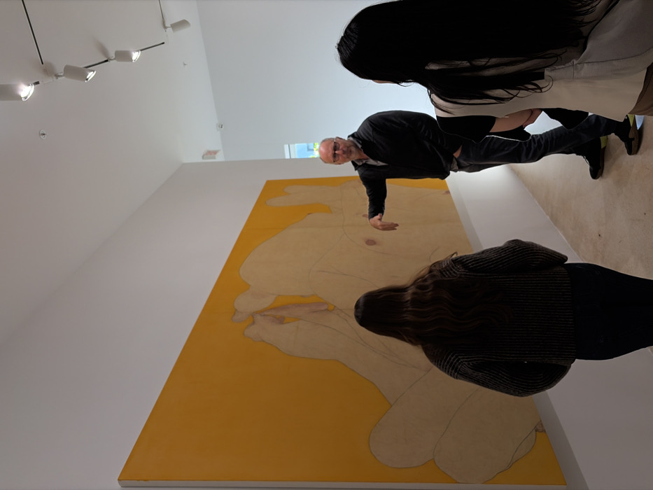 The image depicts an interior scene within a modern art gallery or museum located in Miami, United States of America. The space features pristine white walls, a white ceiling equipped with a black track lighting system holding multiple white cylindrical spotlights, and a light-colored floor.
A large, rectangular painting dominates the left wall. The artwork features a vibrant yellow background upon which large, simplified, light-toned human figures or body parts are outlined with faint lines, some appearing nude with discernible anatomical details.
Three individuals are present. A Caucasian male, appearing middle-aged with short grey hair, a beard, and glasses, stands in the middle-right. He wears a dark long-sleeved shirt, dark trousers, and dark shoes. His right arm is extended, and his hand gestures towards the lower-right section of the large painting, while his left hand rests in his pocket. He is actively speaking or explaining, engaging with the artwork and the other two individuals.
Two women are observing the scene. The woman on the far left stands with her back to the camera, revealing long, dark, wavy hair. She is dressed in a dark, textured long-sleeved top and dark pants. The second woman is partially visible on the far right, facing left towards the male speaker and the artwork. She has long, straight dark hair and wears a light-colored outer garment over a darker layer.
In the background, to the right of the male figure, an open doorway leads into another white-walled area. Through this opening, a window admitting natural light is visible, and a red "EXIT" sign is mounted on the wall above the doorway.