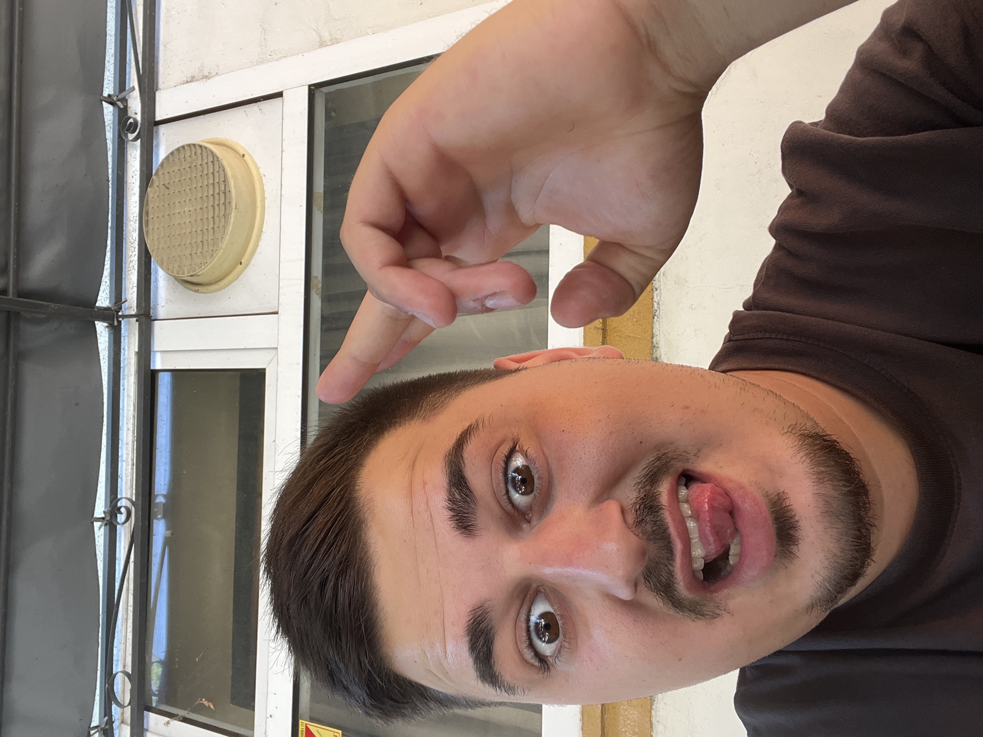 A close-up, waist-up, selfie-style photograph features a young man with dark hair, a mustache, and a goatee. He is wearing a dark brown t-shirt and is positioned in the foreground. His facial expression is animated, with wide-open eyes and his tongue playfully extended. His right hand is raised towards his head, with his index finger extended and bent as if making a gesture.
Behind him, a window frame is visible, with panes that reflect the sky and surrounding environment. Above the window, a grey awning or overhang is present. To the right of the window, a round, textured, light-colored object, possibly a vent cover, is mounted on the wall. The wall itself appears to be painted white and has a yellowish trim along the bottom.
The overall setting suggests an outdoor or semi-outdoor location, likely a public space given the presence of the window and exterior elements. The man's playful demeanor and the selfie format indicate a casual and personal moment. The image does not contain any specific landmarks or elements that would definitively place it in the city of Akandzhievo, Bulgaria.