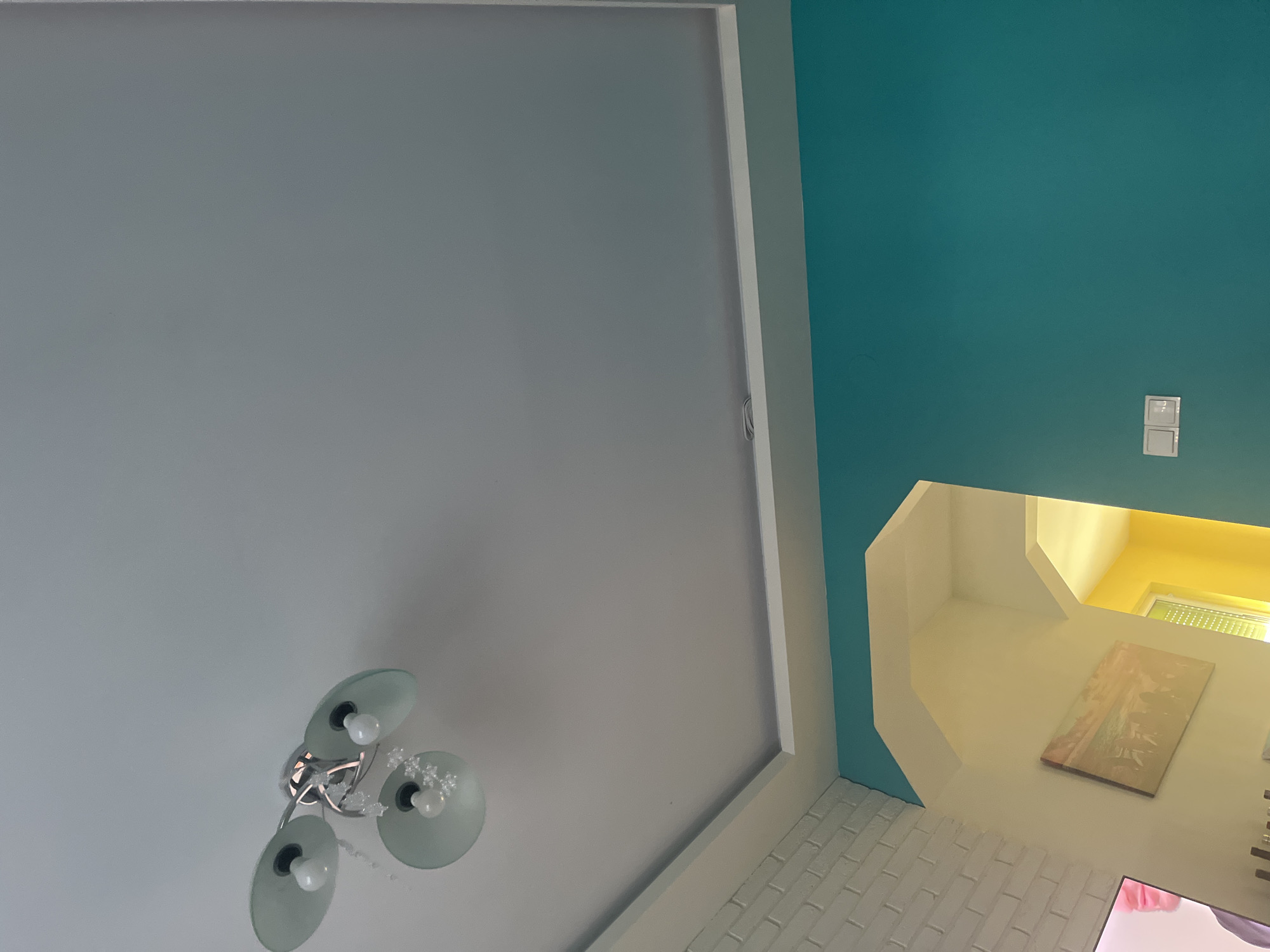 The image shows an interior view of a room, with the camera pointing upwards. The ceiling is primarily gray, with a modern, multi-bulb light fixture hanging from it. The walls are painted in two colors: a teal blue and cream color. A painting is visible on the cream wall and a light switch is seen on the teal wall. The floor appears to be white-painted brick. The scene is likely located indoors and does not contain any people. No context is given for Pokrovnik, Bulgaria.
