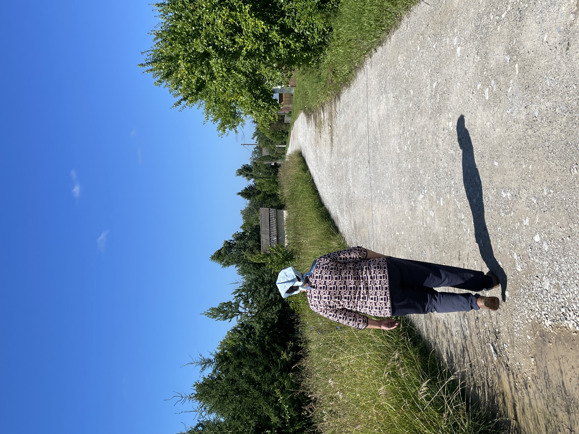 A woman wearing a headscarf and a patterned shirt walks away from the camera down a gravel road in Дресвяново, Russia. The road curves to the right, surrounded by lush green trees and tall grass. In the background, simple wooden houses with dark roofs are visible. The sky is a clear, bright blue with a few wispy white clouds. A shadow of the woman is cast on the road beside her.