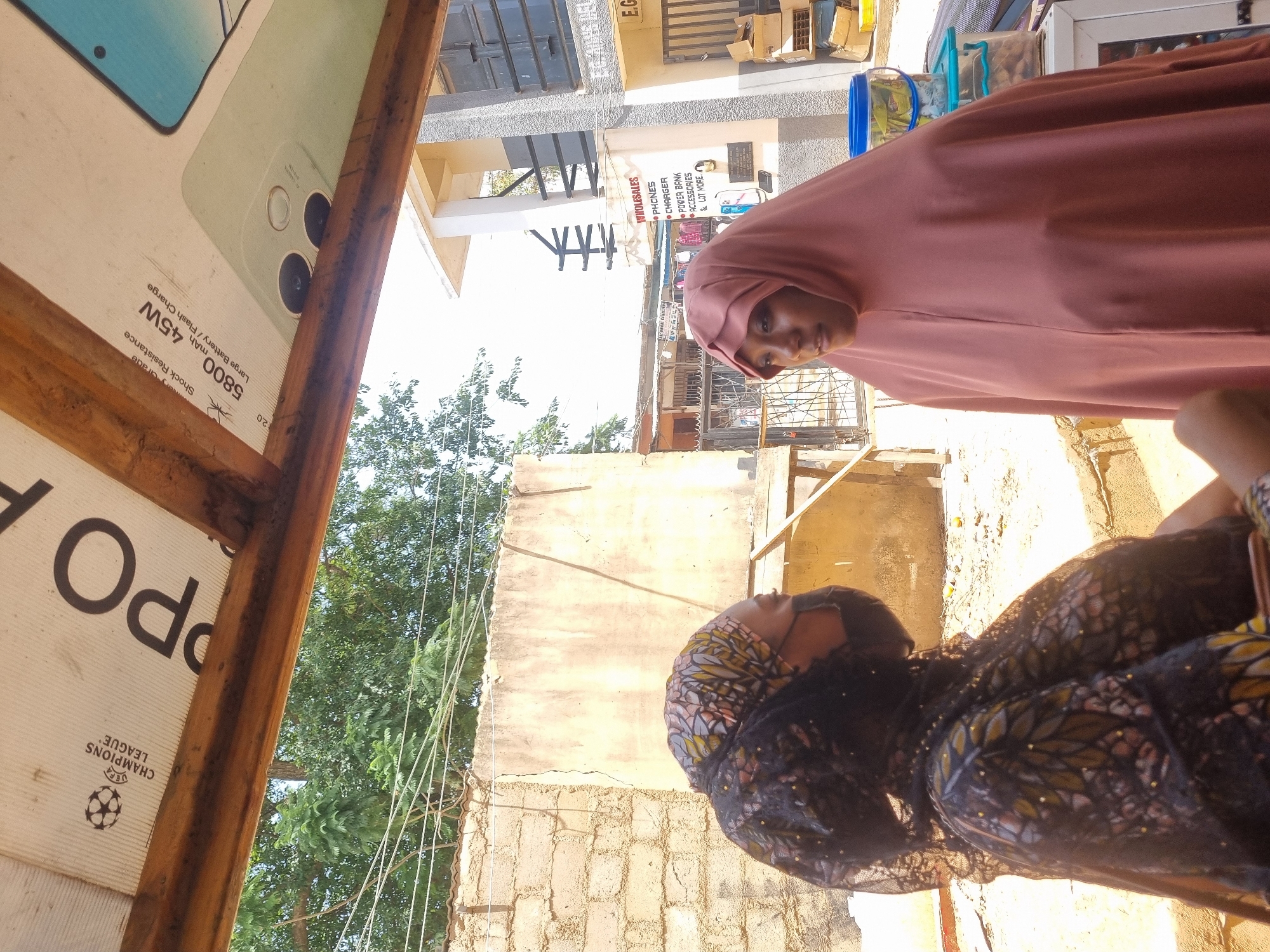 Here is a description of the image:
Two women are visible in this outdoor shot, with a commercial setting in the background. One woman is wearing a hijab in a shade of rose or light brown. The second woman is partially visible, and wearing a dark head covering patterned with gold and brown.
An advertisement for an OPPO mobile phone is partially visible on the left side of the image, with details about the phone's features such as "5800 mAh" and "45W Large Battery/Flash Charge." A sign is visible in the background advertising wholesale phones, chargers, power banks, accessories, and more. The name "E.G. Aminu Stores" is also visible on a sign. The environment appears to be a street scene, with buildings and vegetation visible in the background.