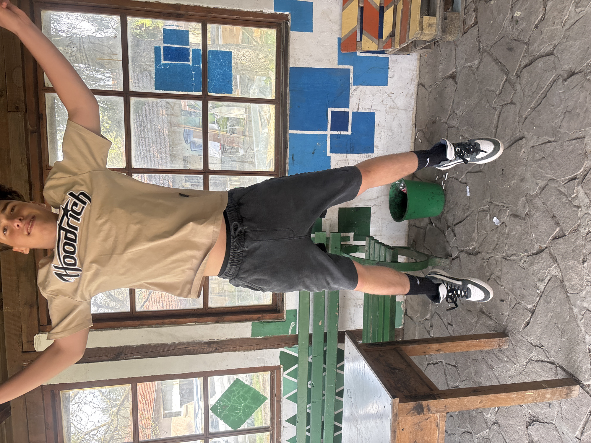 A young person, likely male, is captured in a dynamic pose, appearing to float or jump, with his arms outstretched and legs spread. The image is rotated 90 degrees clockwise, so he appears horizontal against a wall. He is wearing a light brown short-sleeved t-shirt with the word "Hoodrich" printed in black and white on the front, dark grey athletic shorts, black socks with a white Nike swoosh logo, and black and white sneakers. He has short dark hair and a slight smile on his face.
The scene is an outdoor or semi-outdoor space in Troyan, Bulgaria, possibly a patio or covered area. The background features a white wall adorned with an abstract, geometric pattern of various shades of blue squares and rectangles. To the upper right, there are also wooden slats painted in yellow, orange, and red hues, forming another decorative pattern. Several multi-pane windows with rustic wooden frames are set into the walls, revealing glimpses of trees and natural light outside. The ground is paved with irregular, dark grey stone slabs.
In the mid-ground, a green metal bench or railing is visible, and part of a wooden table or structure is in the foreground. A green barrel or trash can is positioned near the person's right foot (when viewed upright), with small white debris scattered on the ground around it. The lighting suggests it is daytime with bright, natural light, and the overall mood is casual and playful.